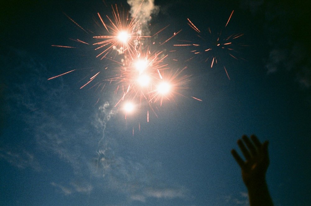 A hand is visible in the lower right corner of the image, the hand that has just released a small firecracker. Red light explodes in the top left and center of the image. Behind both the hand and fireworks are a dark blue sky with clouds and smoke