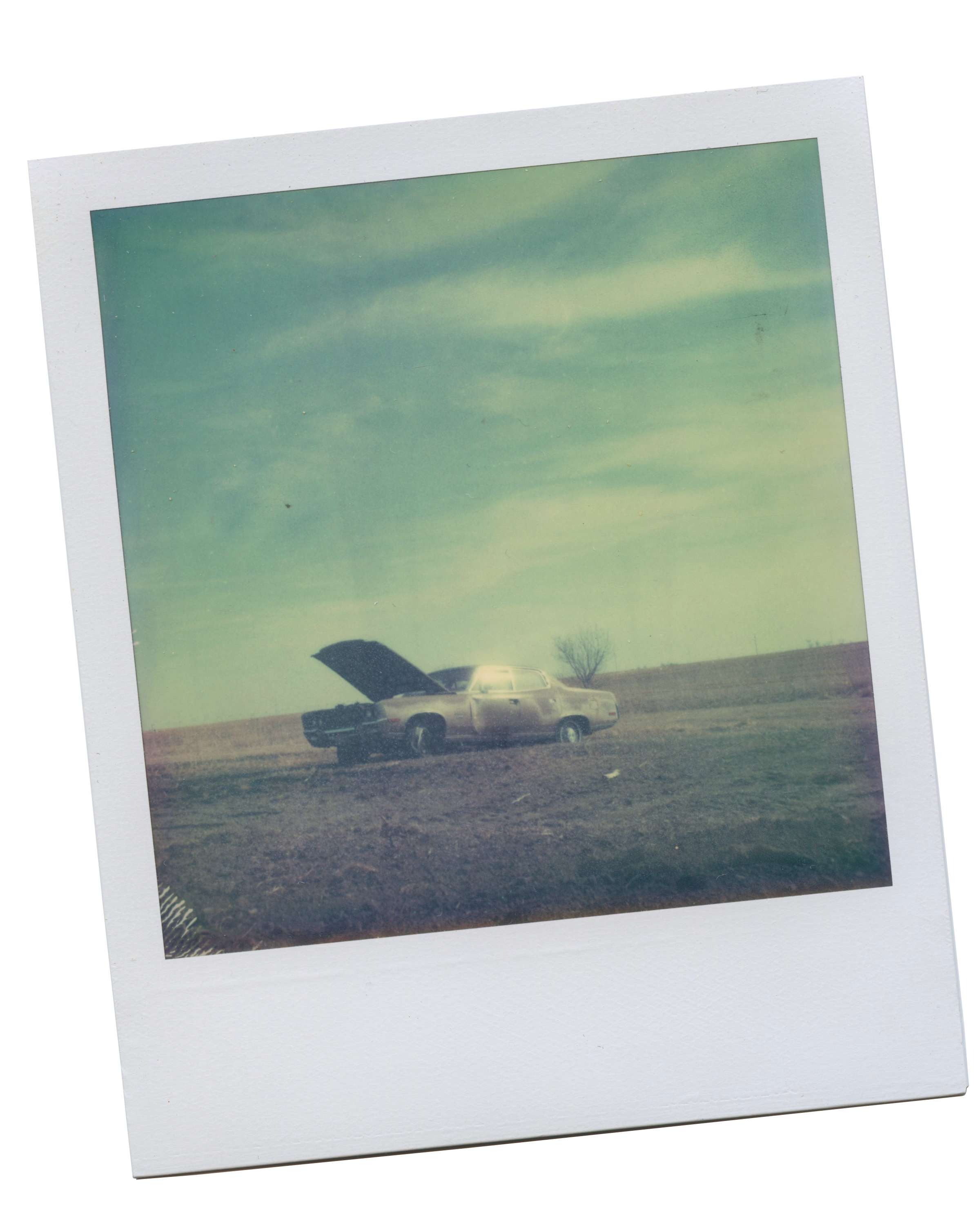 polaroid of a car in the middle of an empty brown field. The hood is up, and the sun is reflecting off of the window.