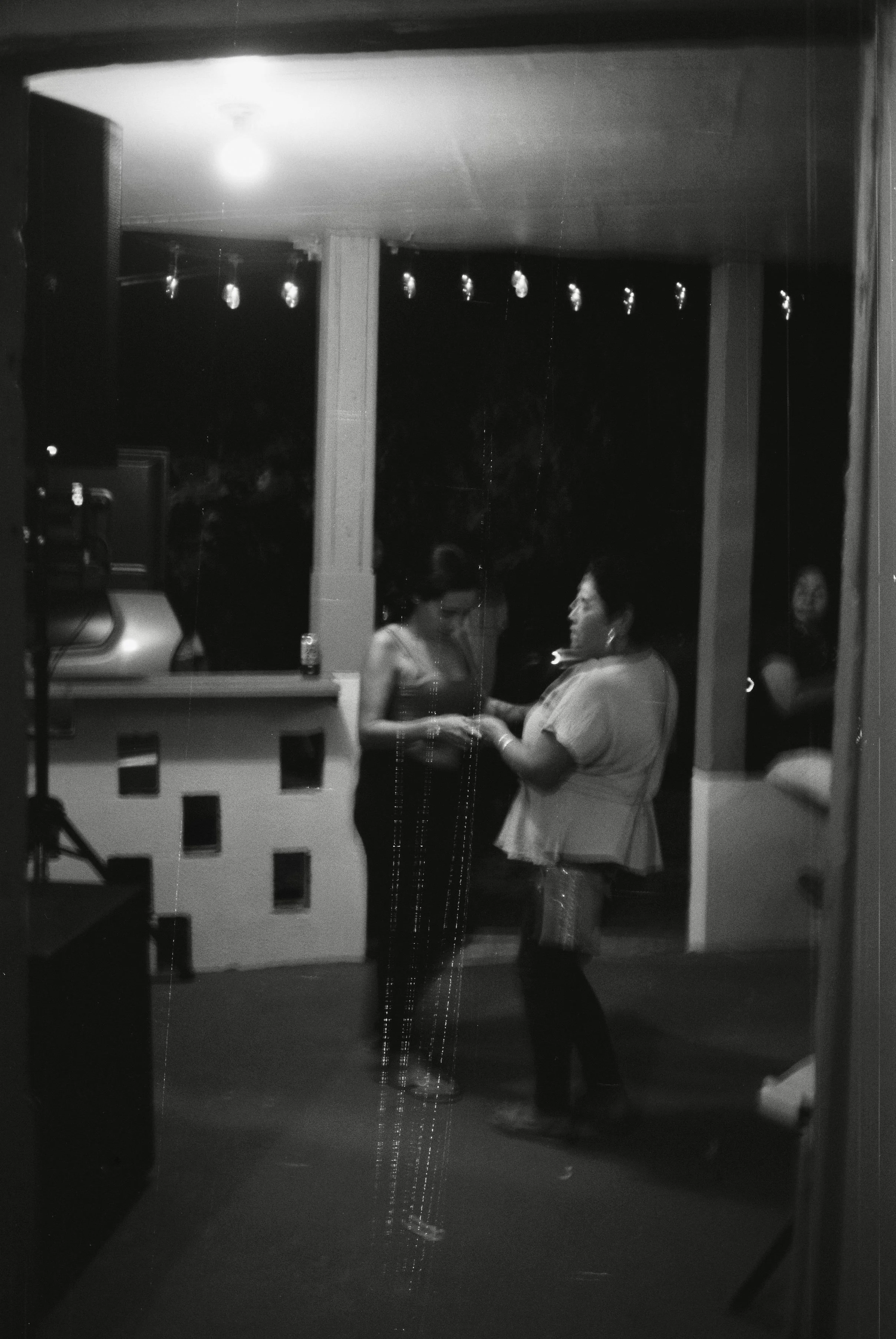 Jannette and her daughter dance to the music playing on a DJ set-up to the left of the frame. They are slightly blurry in the low light. The glitter of Jannette’s earrings and purse, and the Christmas lights decorating create trails of movement.