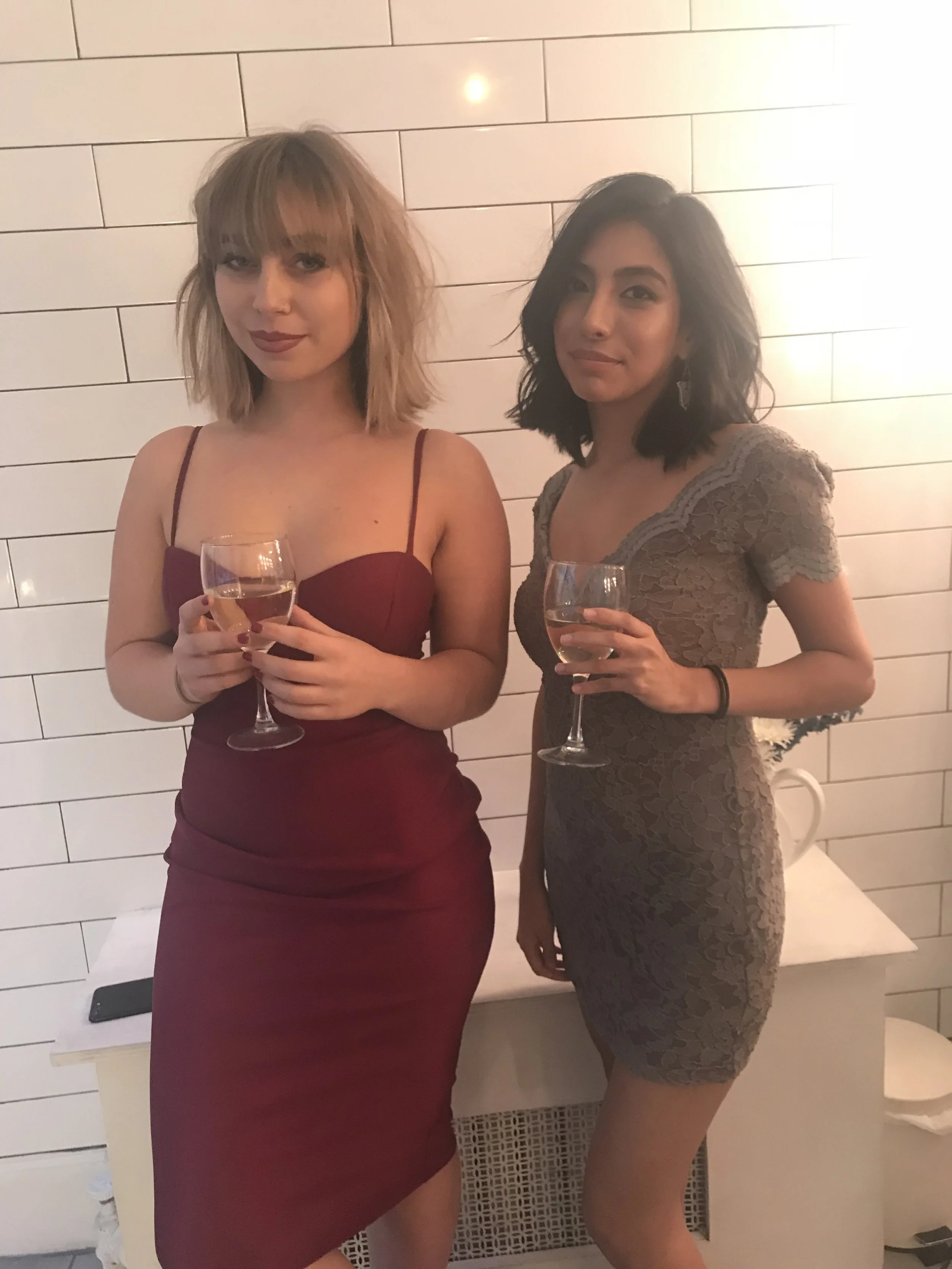 Two women dressed in elegant dresses holding wine glasses, standing in front of a tiled wall at a social event.