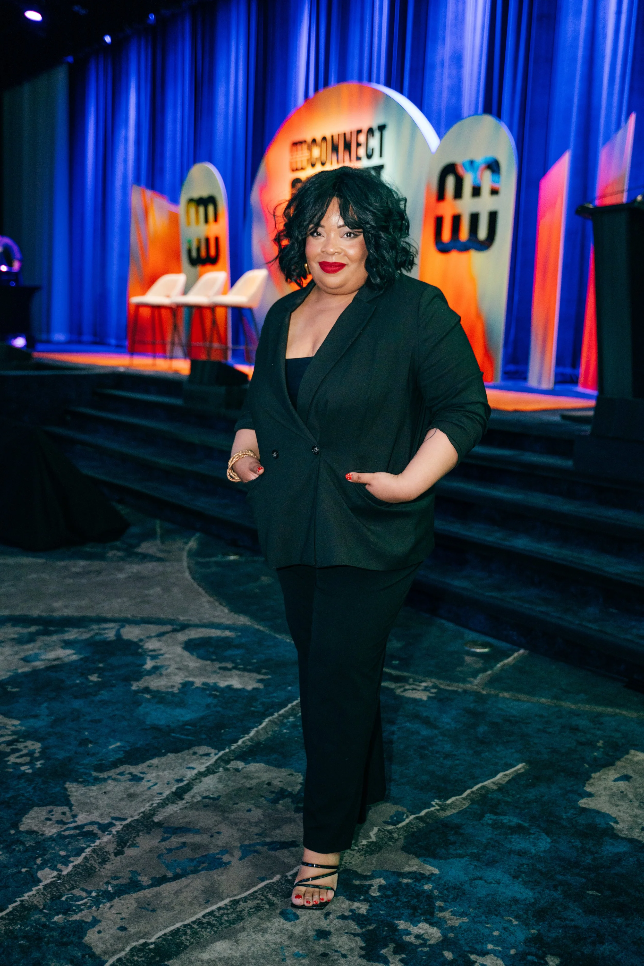 A woman with curly black hair and red lipstick wearing a black blazer and black pants, standing on a patterned carpet in front of a stage with blue curtains and colorful decorative panels.