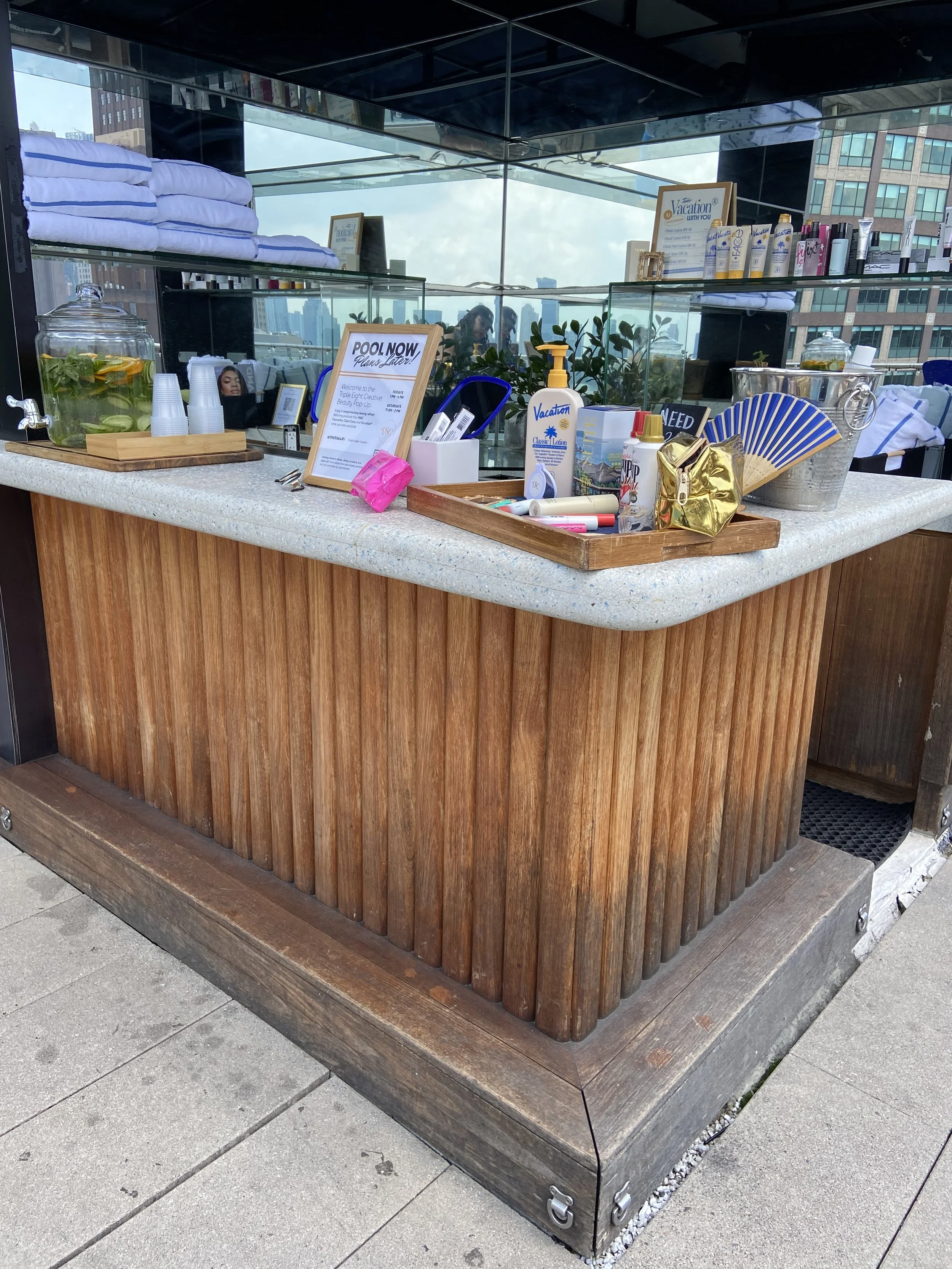 A rooftop bar or lounge with a wooden and glass counter, showing towels, bottles of lotion, and promotional materials, with city buildings in the background.