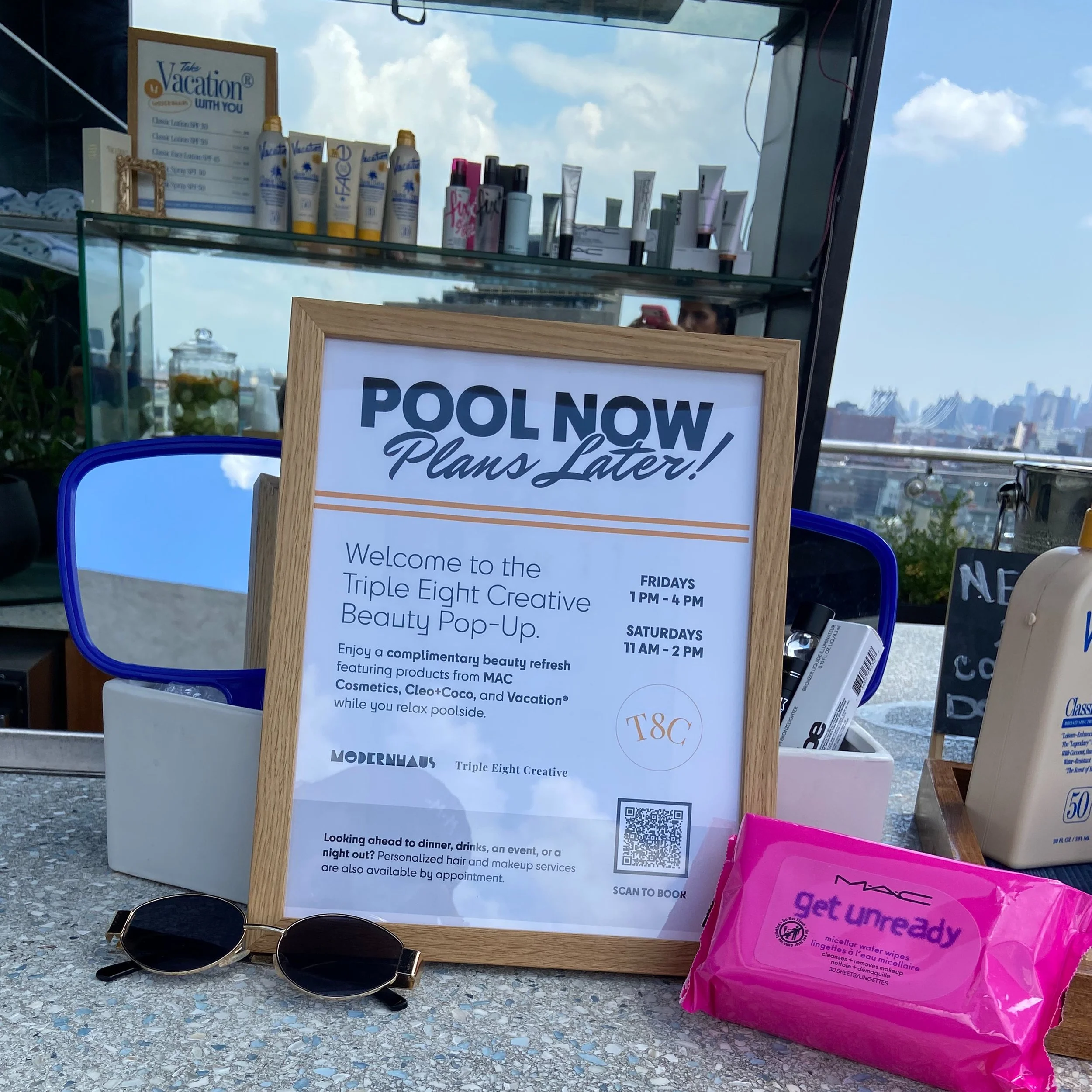 A promotional display at an outdoor event showing a sign for a beauty event called 'Pools Now, Plans Later' with details about the event happening Fridays and Saturdays, offering complimentary beauty refreshments and services such as hair and makeup. The display includes a glass case with skincare products, a pair of sunglasses, a pink bag, and some other smaller items, with a city skyline and cloudy sky in the background.