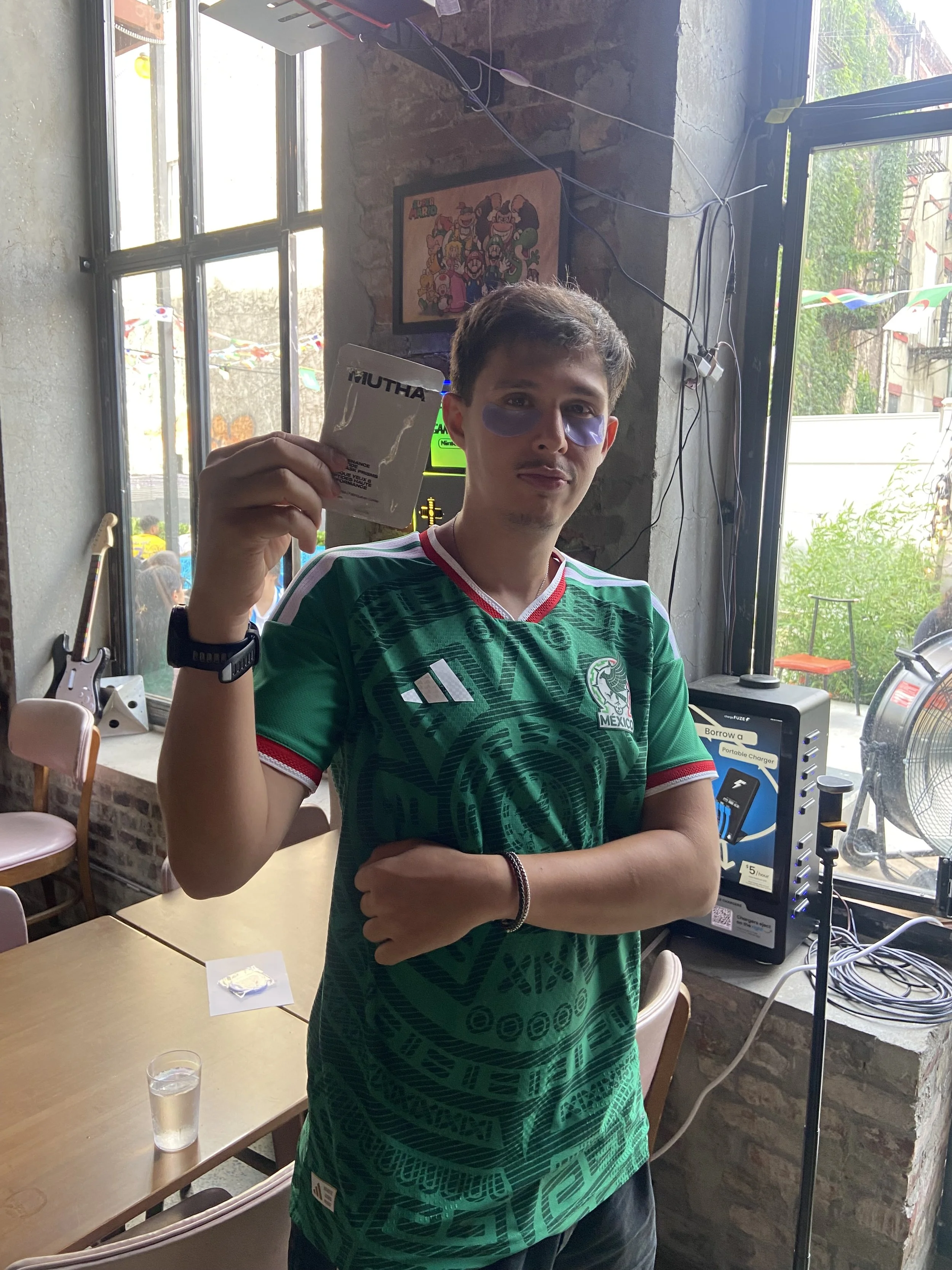 A young man with short brown hair wearing a green Mexican soccer jersey, purple eye masks, and black wristbands, standing indoors near a window and holding a packaged item in his right hand, with various items and a framed cartoon poster on the wall behind him.