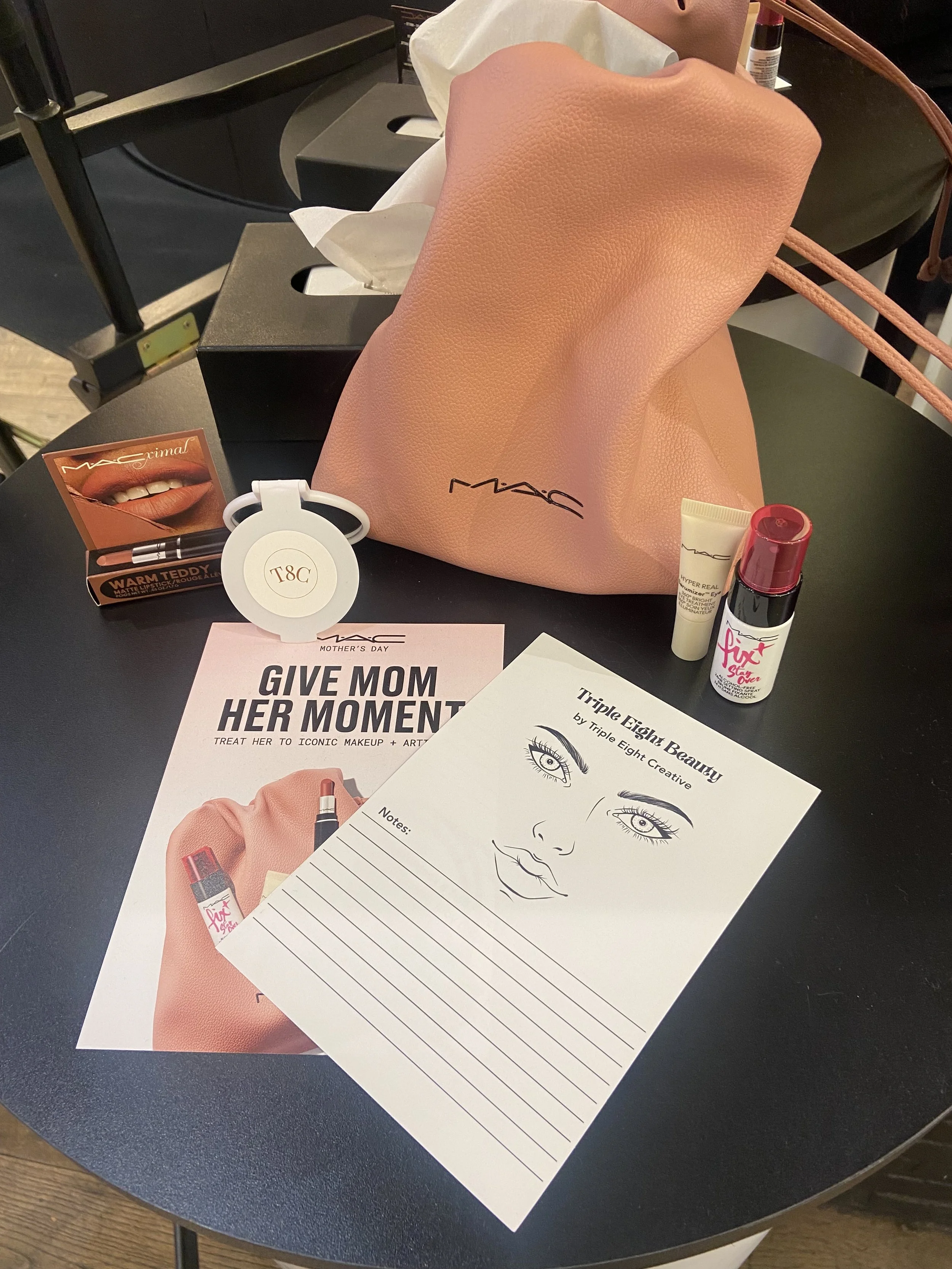 A table with promotional materials, including a MAC cosmetics bag, a makeup guide with eye illustrations, and beauty products such as lipstick and face cream. The materials celebrate Mother's Day with the message "Give Mom Her Moment."