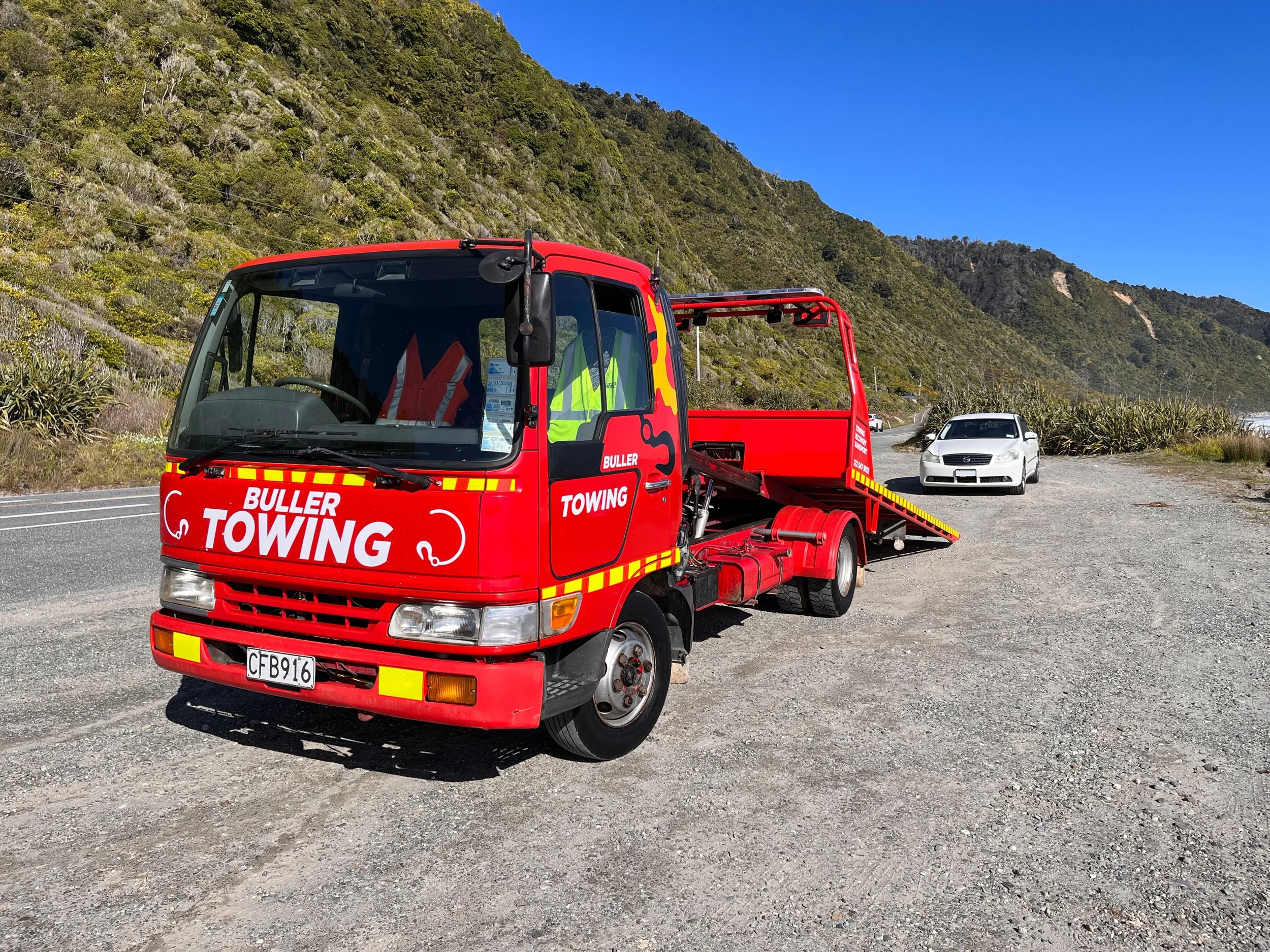 The distinctive Buller Towing & Recovery towing truck parked on a gravel roadside, with a white car behind it, against a backdrop of green hillside and clear blue sky on the Coast Road