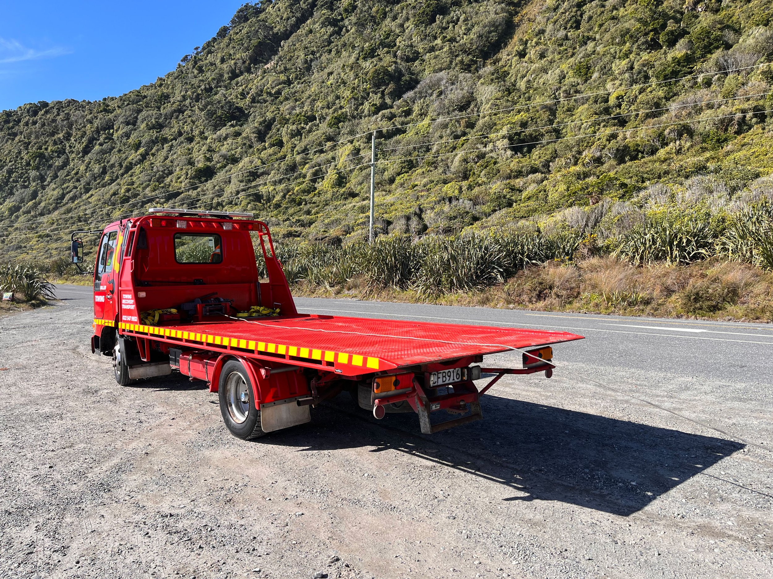 The Buller Towing & Recovery flatbed tow truck parked on the side of the Coast Road with a scenic hillside covered in bushes and trees in the background
