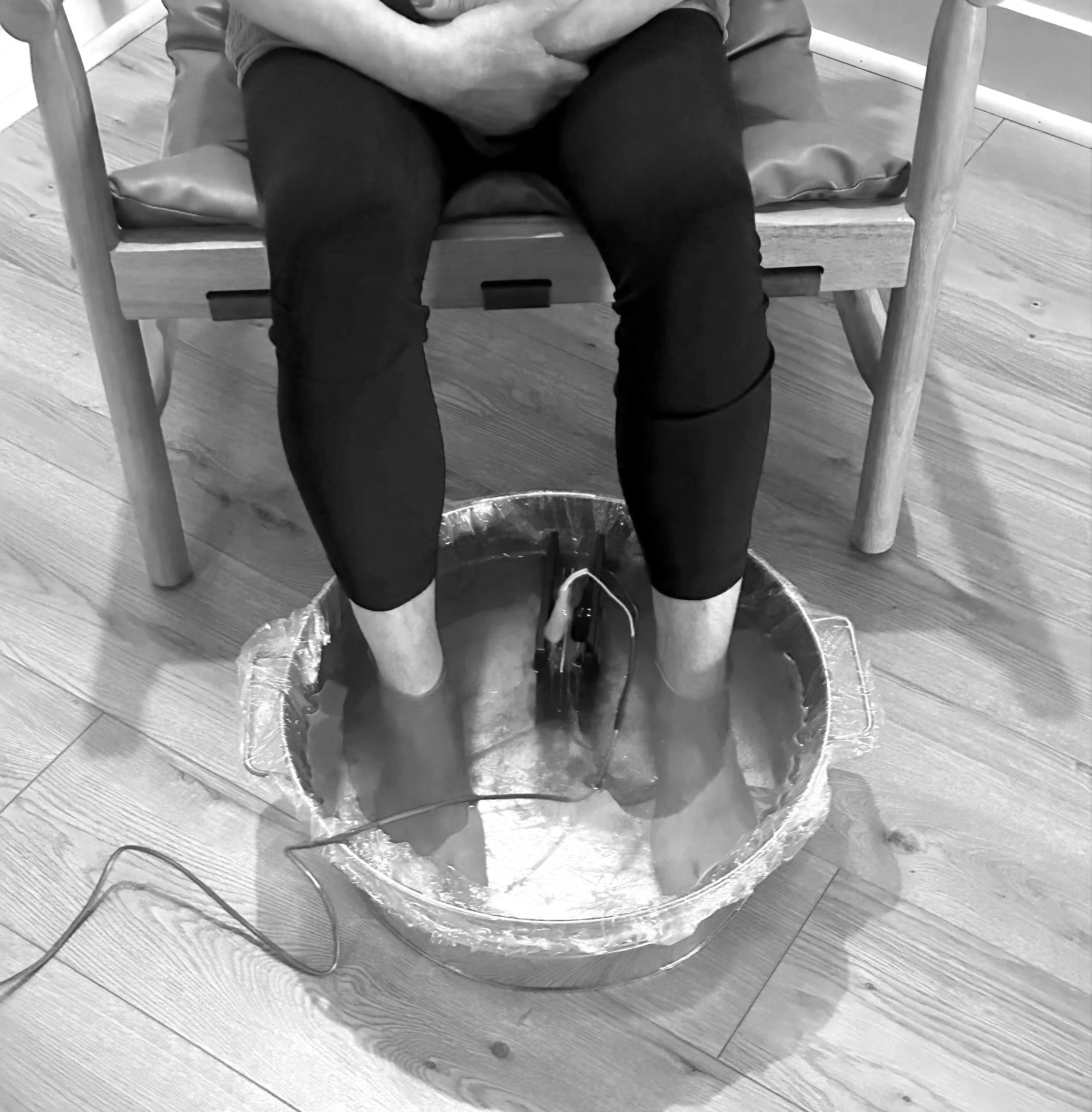 Person soaking their feet in a basin on the floor, sitting on a wooden chair, with their legs submerged in water.