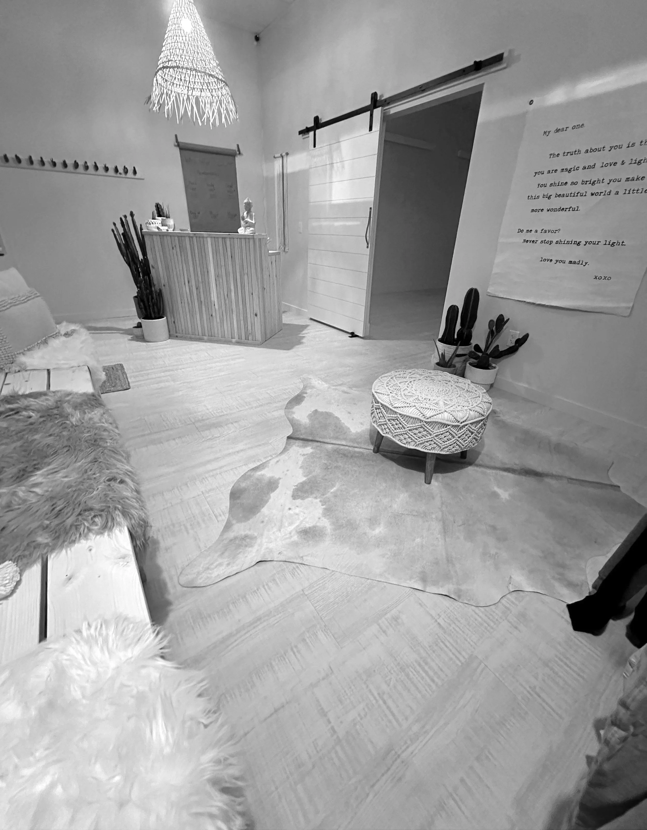 A cozy interior room featuring a plush cowhide rug, a small round stool with a woven pattern, a bench with fur and blanket, potted cacti, a sliding barn door, and decorative wall hangings.