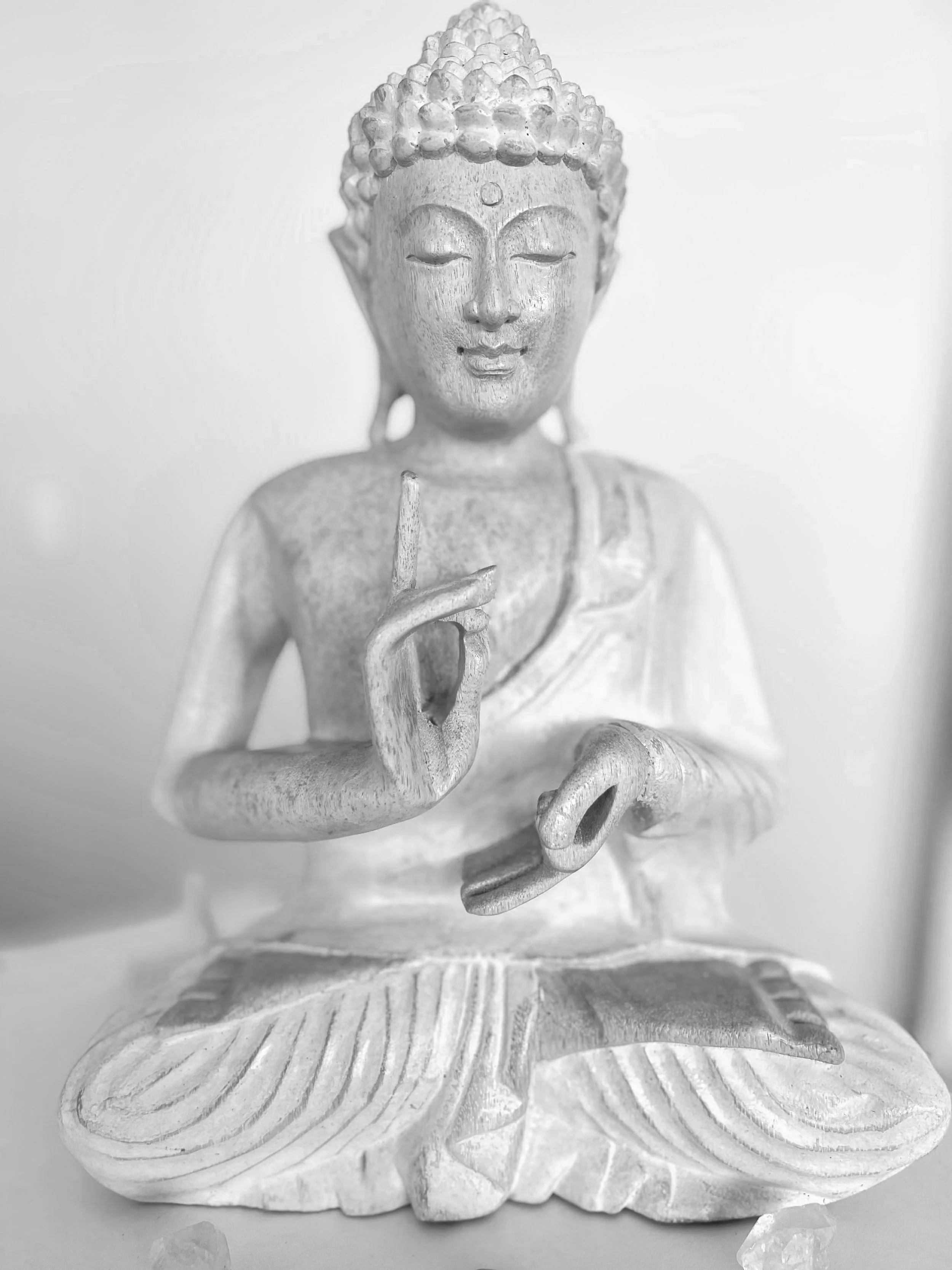 A white Buddha statue in a seated meditative pose with eyes closed, holding a small object between the thumb and index finger of the right hand, and resting the left hand on the lap with fingers pointed downward.