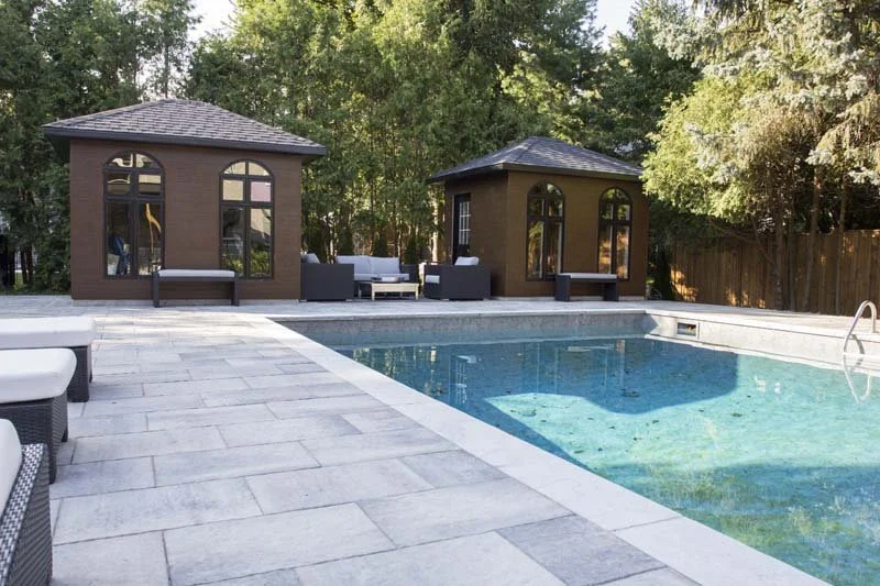 Backyard with a swimming pool, two small wooden cabanas, outdoor seating, and trees in the background.