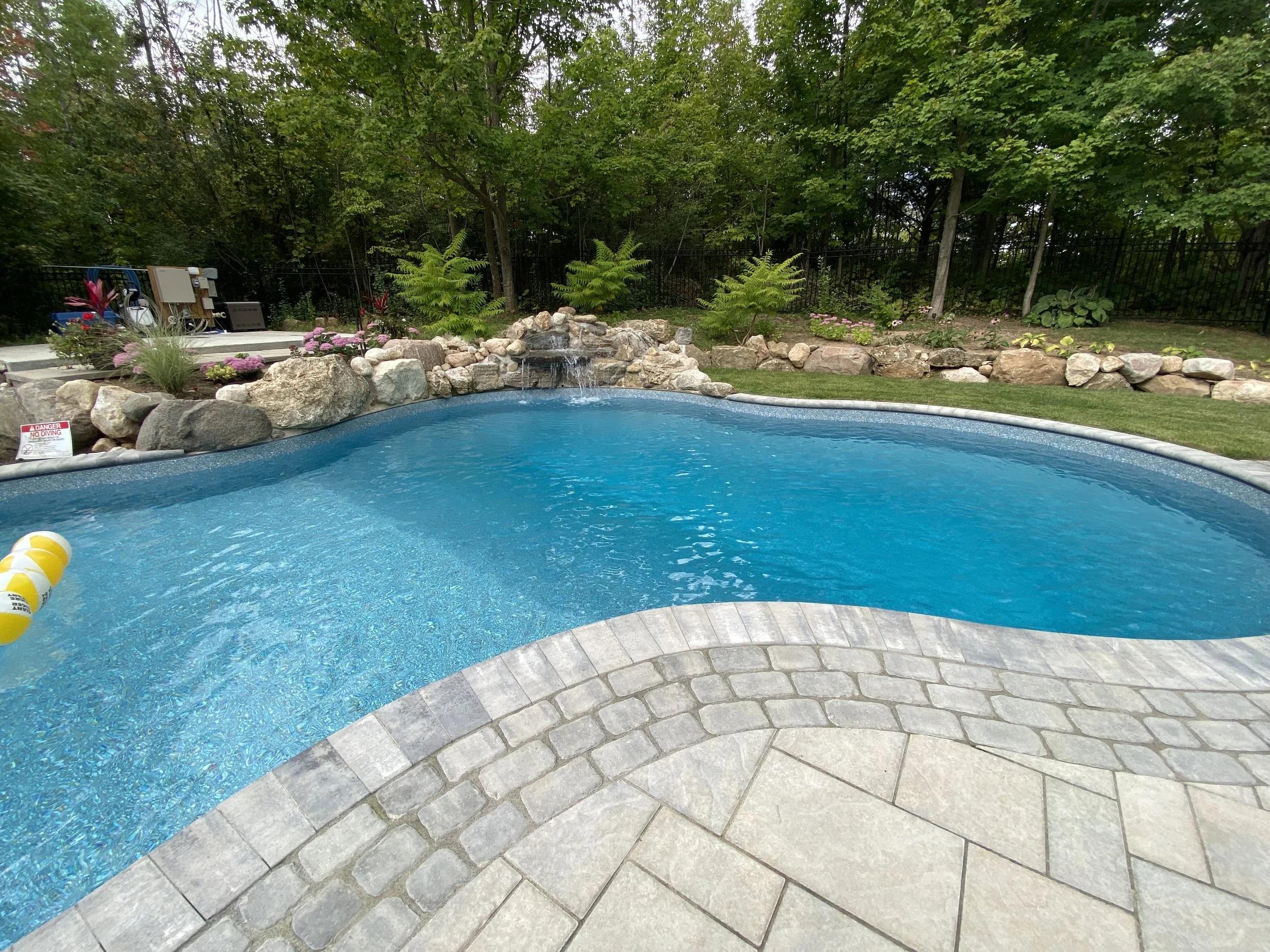 Backyard swimming pool with clear blue water, surrounded by stone pavers and rocks with a small waterfall feature, lush green trees and plants in the background.