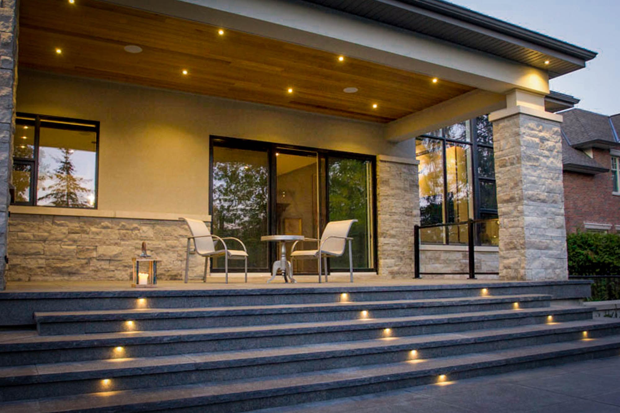Luxury home's covered patio area with two chairs and a small table, illuminated by embedded lighting on steps and ceiling, with stone and glass exterior walls.
