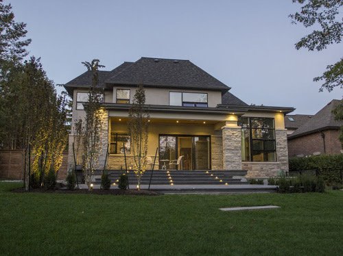 Modern two-story house with a dark roof, stone and stucco exterior, large glass windows, and a well-maintained lawn at dusk.
