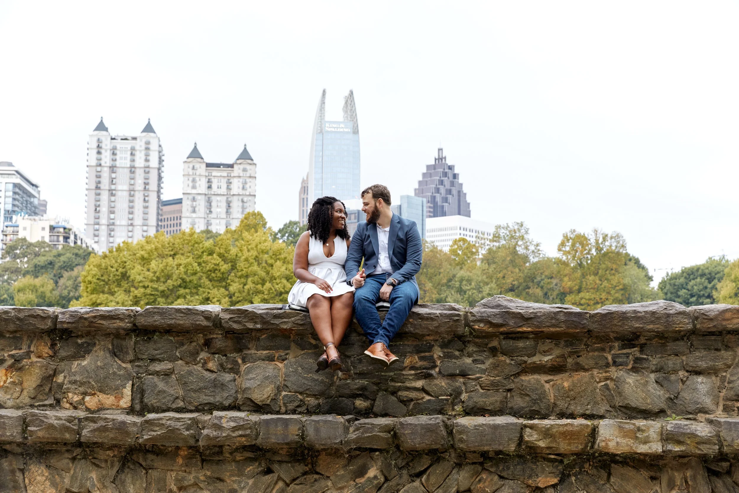 Engaged couple sitting on a brick wall in front of the Atlanta skyline