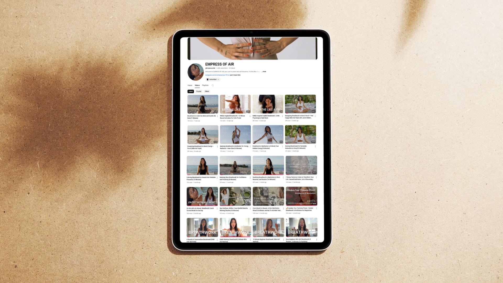 A tablet displaying a YouTube channel page named EMPRESS OF AIR with various videos related to fitness, yoga, and relaxation, placed on a textured beige surface.
