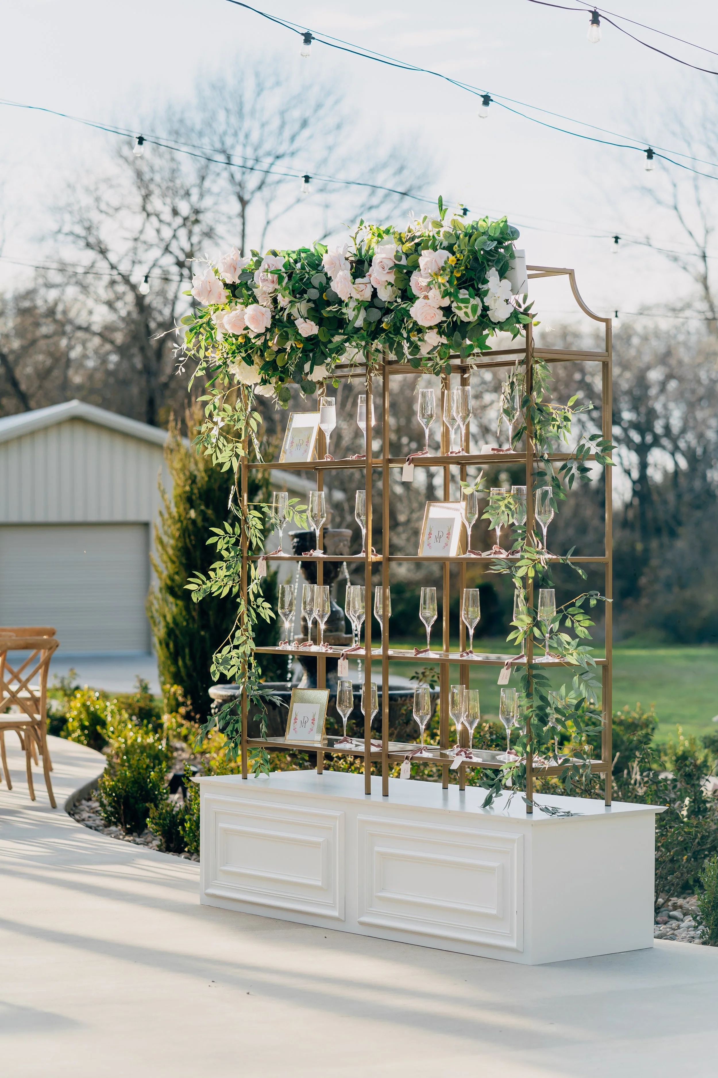 Outdoor wedding cocktail hour with gold shelves and a floral display with white and pink flowers, greenery.