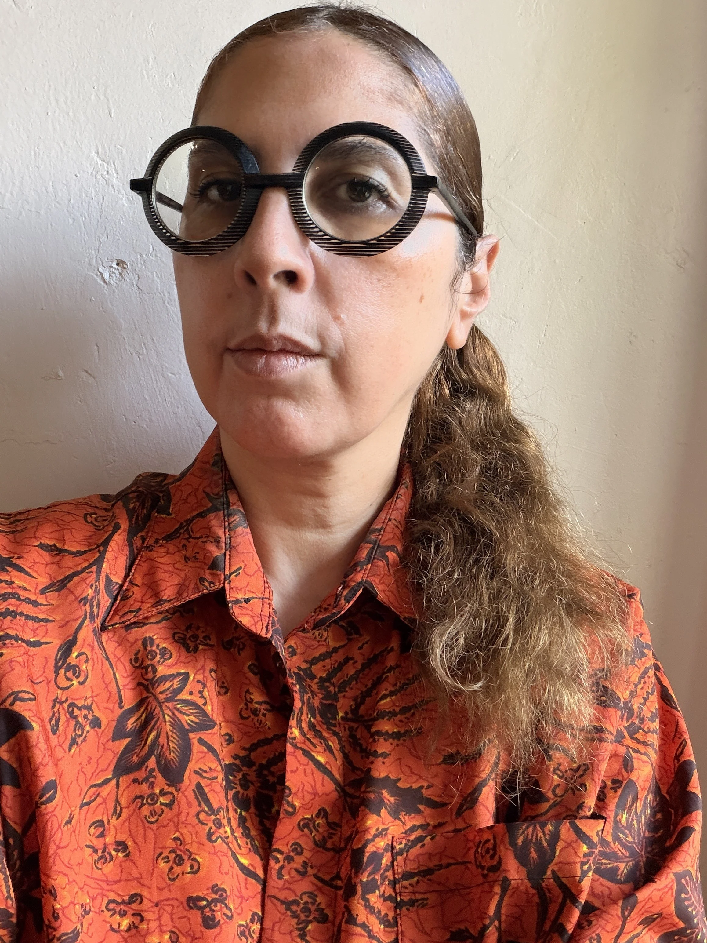 A woman wearing black and white striped round glasses, a red patterned button-up shirt, and has long curly hair pulled over her shoulder, standing against a plain light-colored wall.