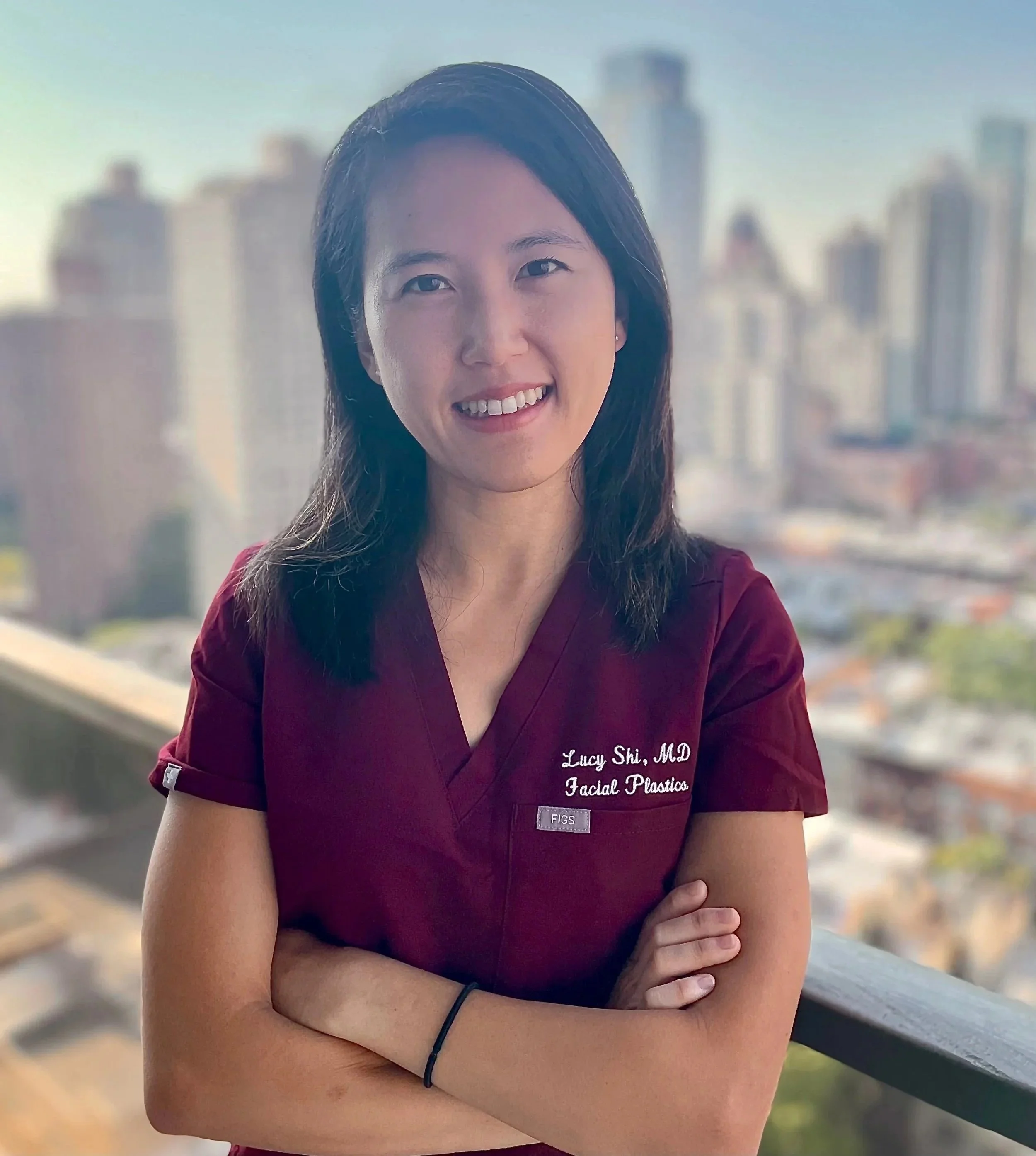 A woman in a maroon medical uniform with the embroidered text 'Lucy Shi, MD Facial Plastics' standing with arms crossed on a balcony overlooking a cityscape with tall buildings and a clear sky on the Upper East Side of New York City's Manhattan.