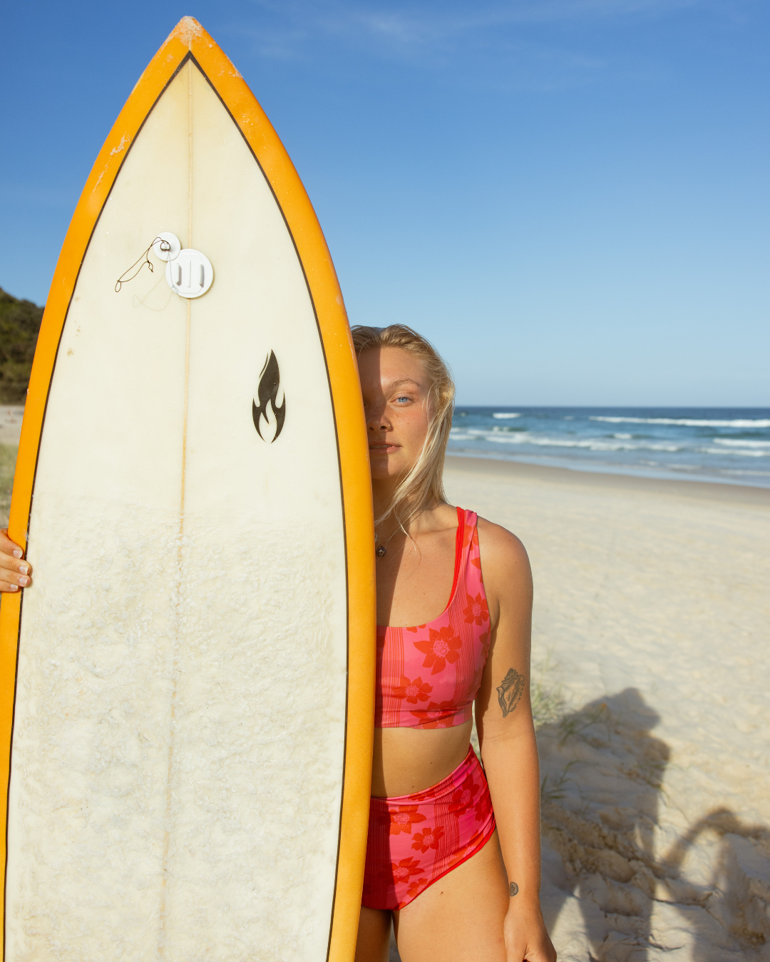 A woman in a pink floral swimsuit holding a surfboard on a sandy beach with the ocean in the background.