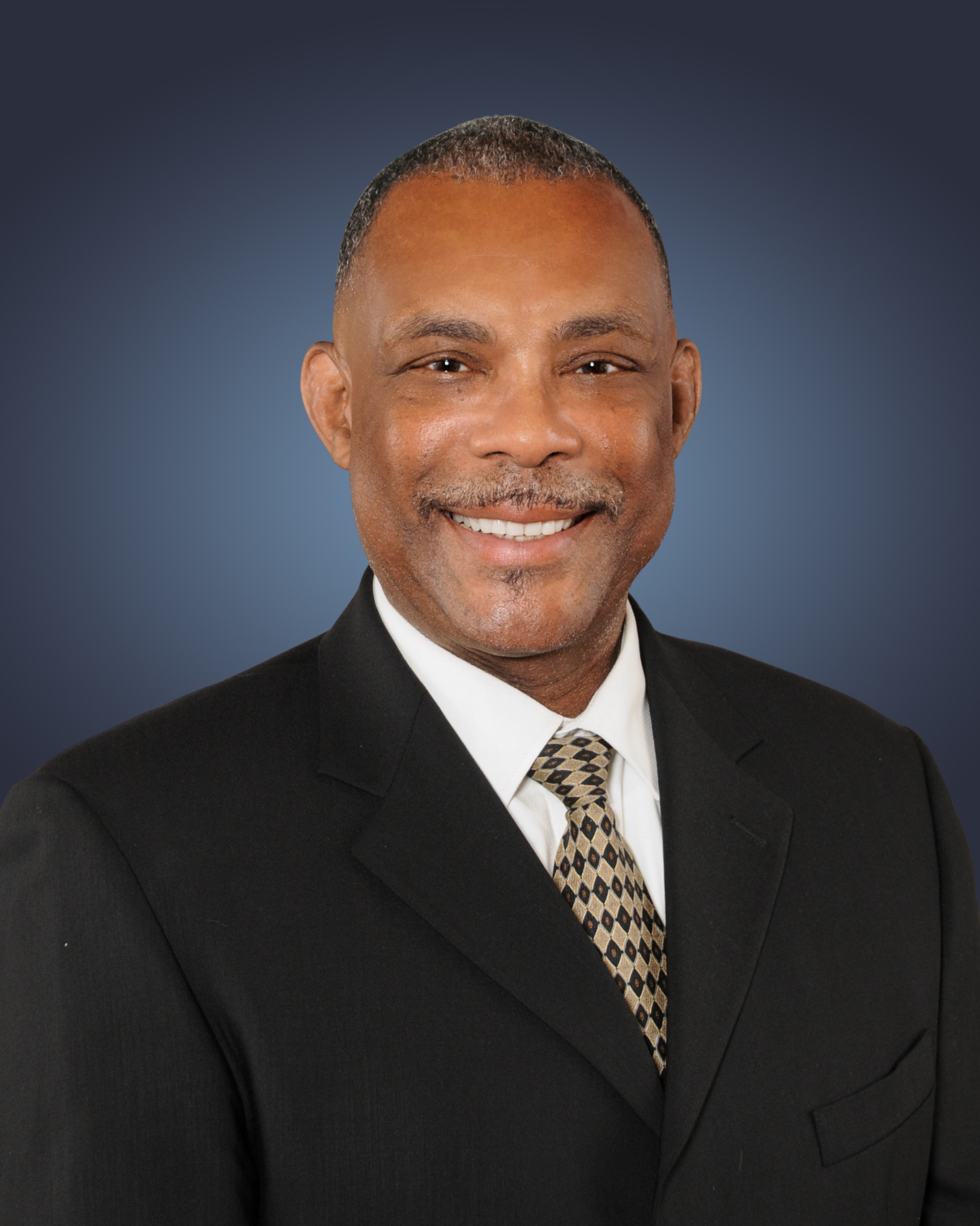 Professional headshot of Tyrone Aiden in a gray suit, white shirt, and patterned tie against a dark gradient background.