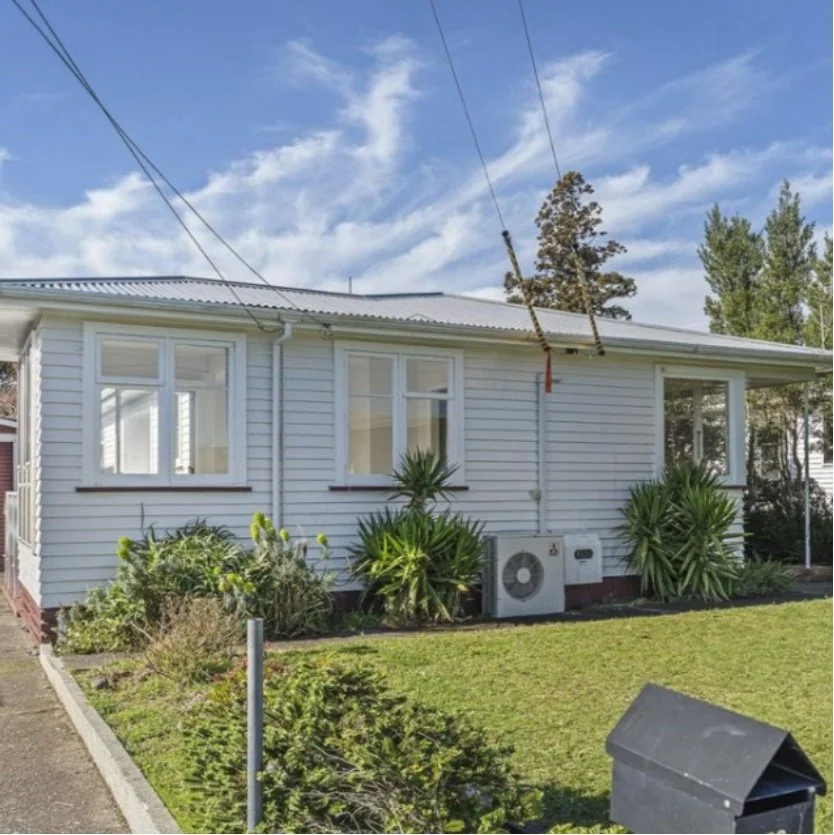 A white house with three large windows, surrounded by green plants and bushes, with a grassy lawn in front. The sky is blue with some clouds, and there are power lines overhead. Wolfe Property client investment property