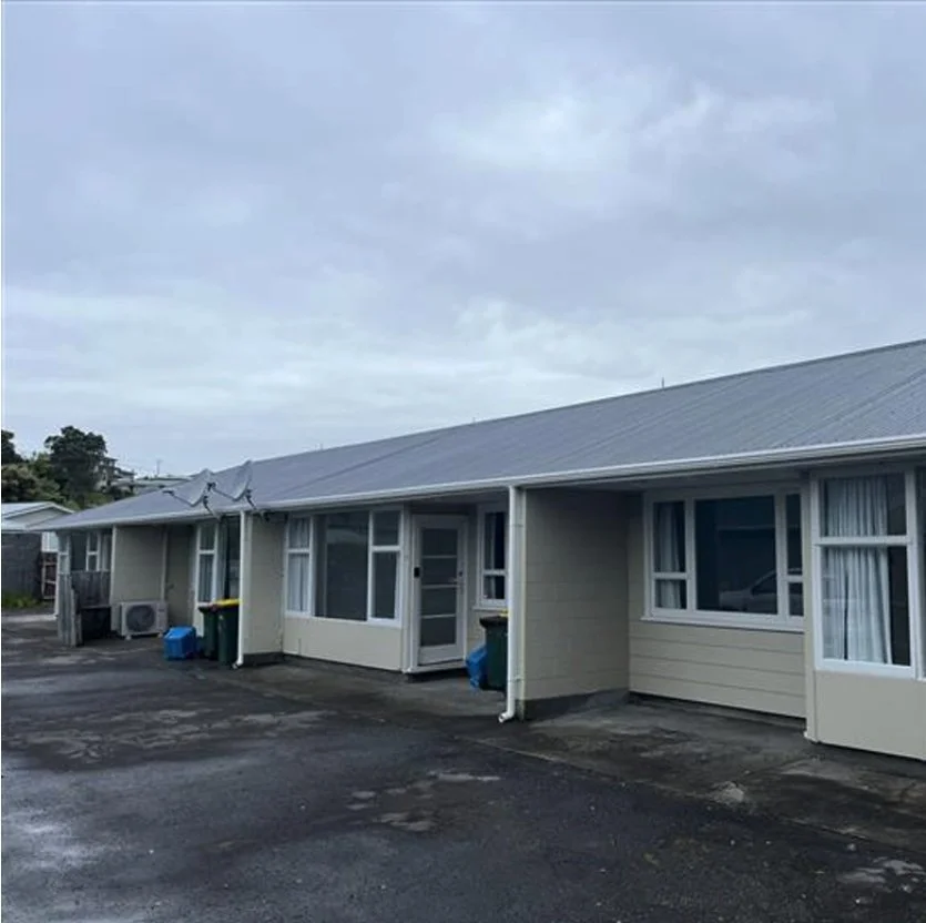 A row of single-story residential townhouses with beige siding and white trim, under a cloudy sky, with a paved parking area in front. Wolfe Property client investment property