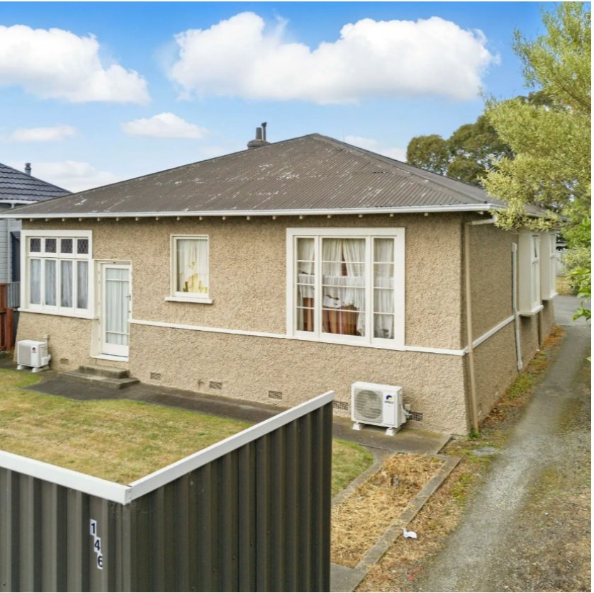 Single-story house with beige stucco exterior, large front window, side window, and white door, with a small yard, air conditioning units, and a gravel pathway. Wolfe Property client investment property.