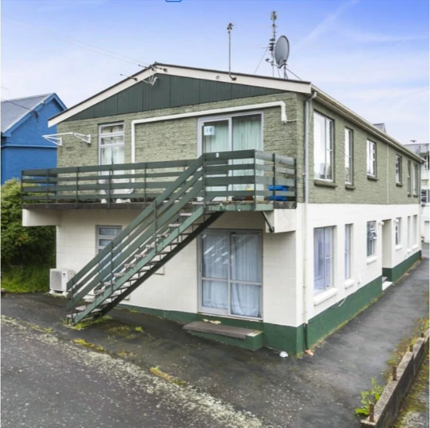Two-story house with exterior staircase, green and white exterior walls, satellite dish on roof, and multiple windows. Wolfe Property client investment property