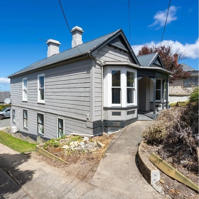 A two-story house with gray wooden siding, white trim, and a metal roof, featuring a small porch and several windows, under a blue sky with scattered clouds. Wolfe Property client investment property.