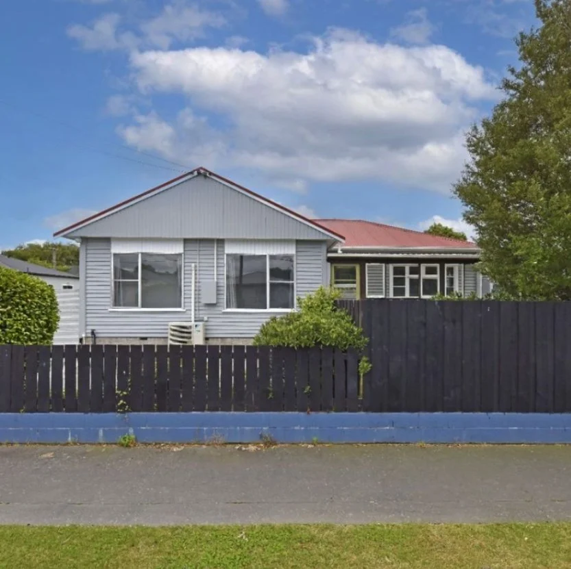 A house with gray siding and a red roof, surrounded by a black wooden fence, with a sidewalk in front and trees on the right side, under a partly cloudy sky. Wolfe Property client investment property.