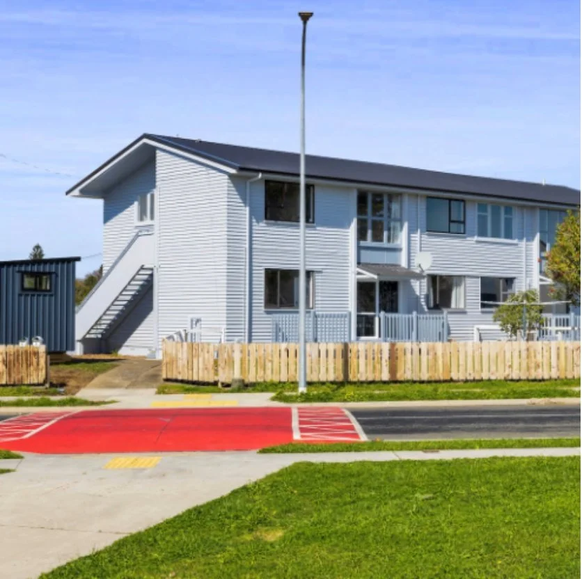 White multi-story residential building with stairs on the side, surrounded by a wooden fence, next to a sidewalk with a red curb and green grass, under a blue sky. Wolfe Property client investment property.