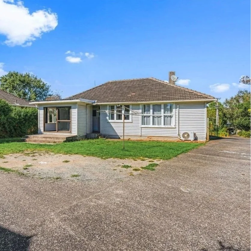 A single-story house with a sloped roof, gray siding, and a small porch area with steps, surrounded by a concrete driveway and a grassy yard, under a bright blue sky with a few clouds. Wolfe Property client investment property