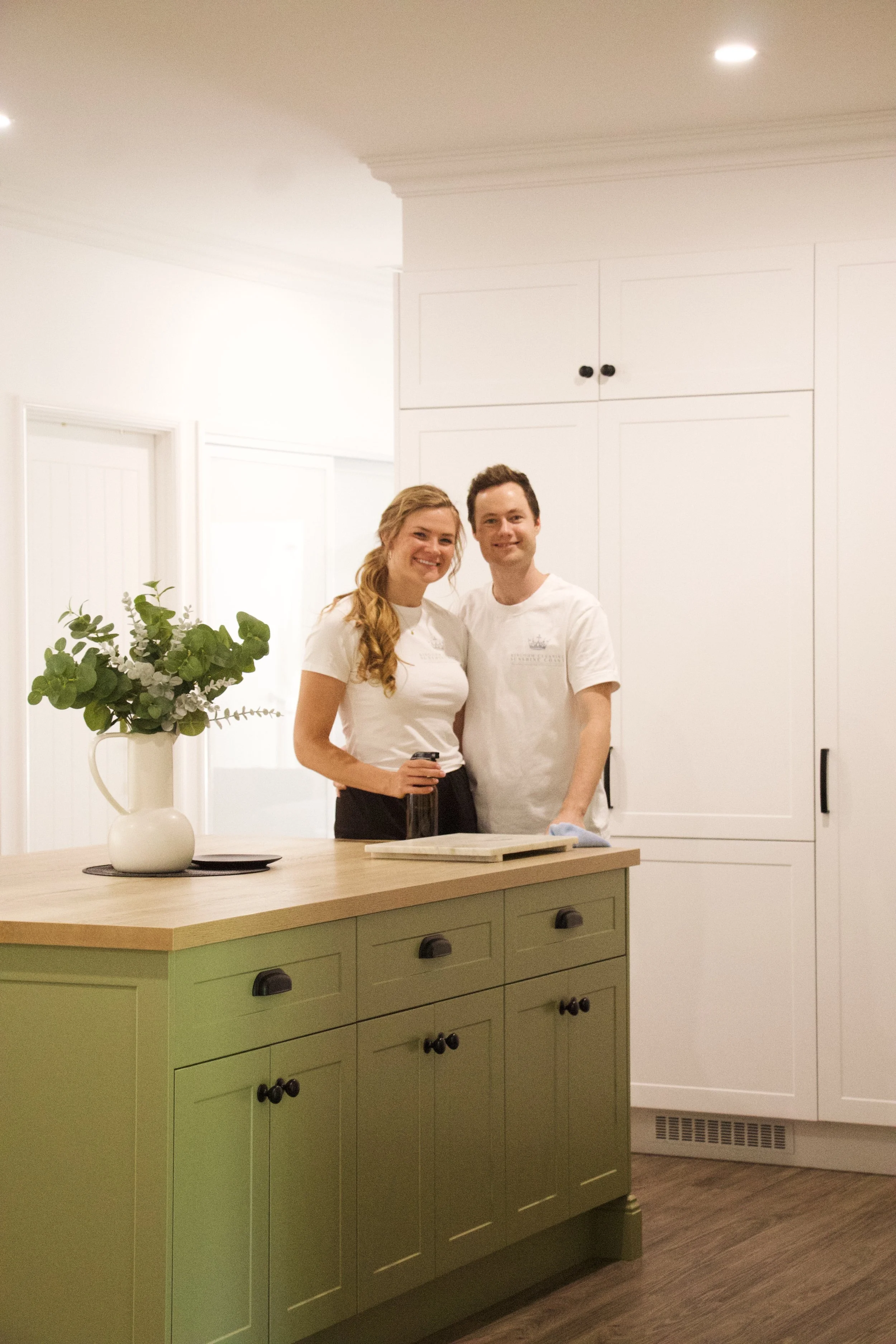 Two smiling people, a woman and a man, standing in a modern kitchen with white cabinets, light-colored walls, a green kitchen island, and a vase with green and purple leaves on the island. Modern home on the Sunshine Coast.