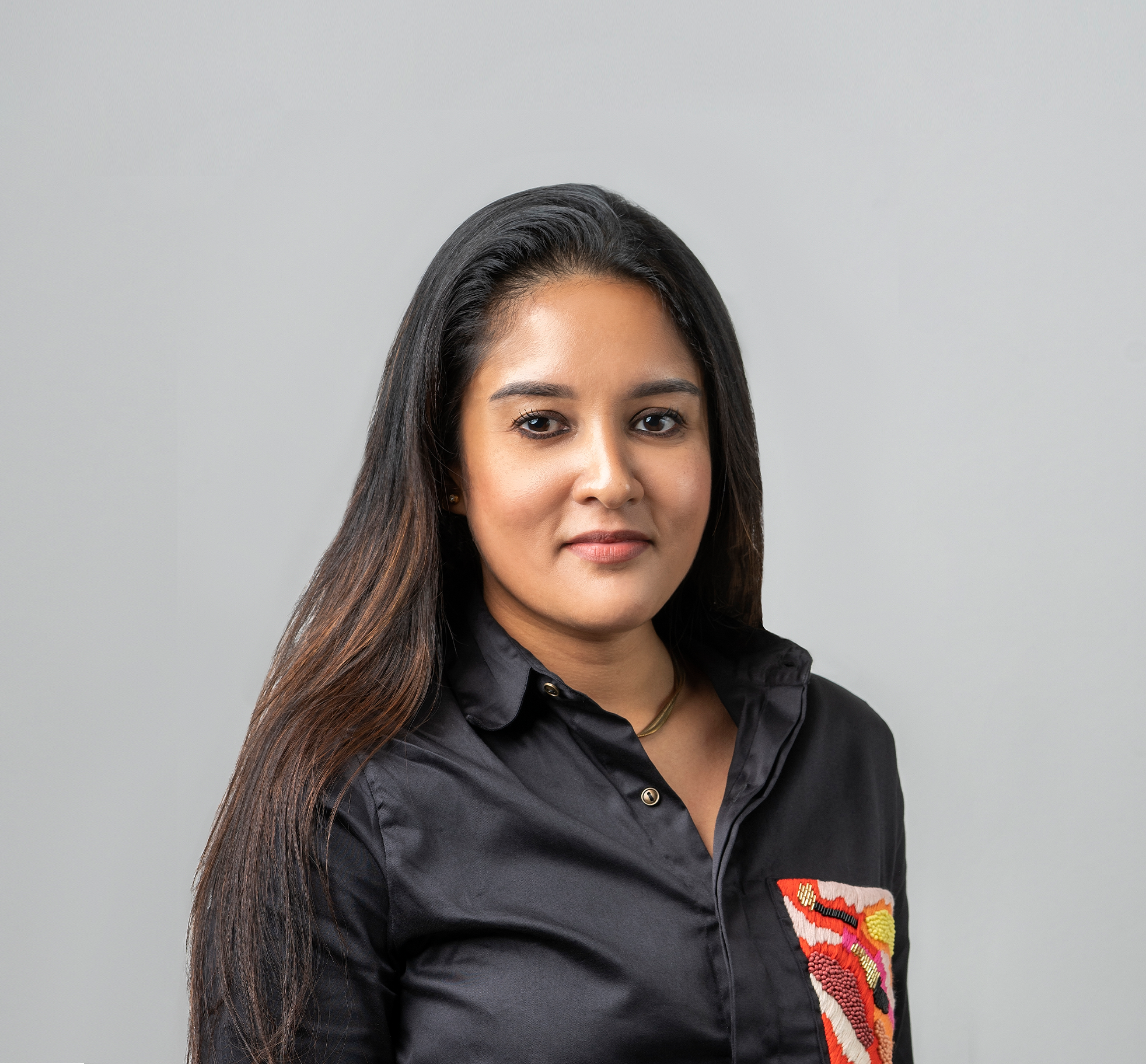 A woman with long dark hair wearing a black collared shirt with a colorful embroidered patch on the chest, standing against a plain light gray background.
