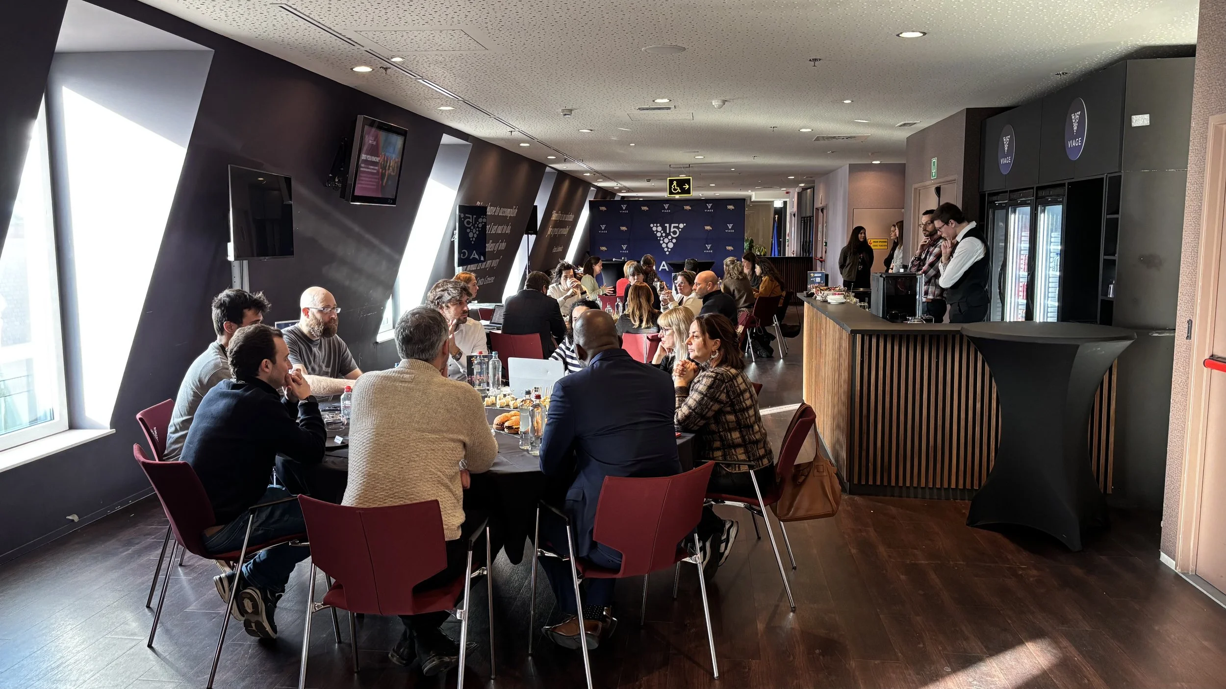 A group of people sitting around a table in a conference room, with some standing by a bar or service counter on the right, and a blue backdrop with logos at the back of the room.
