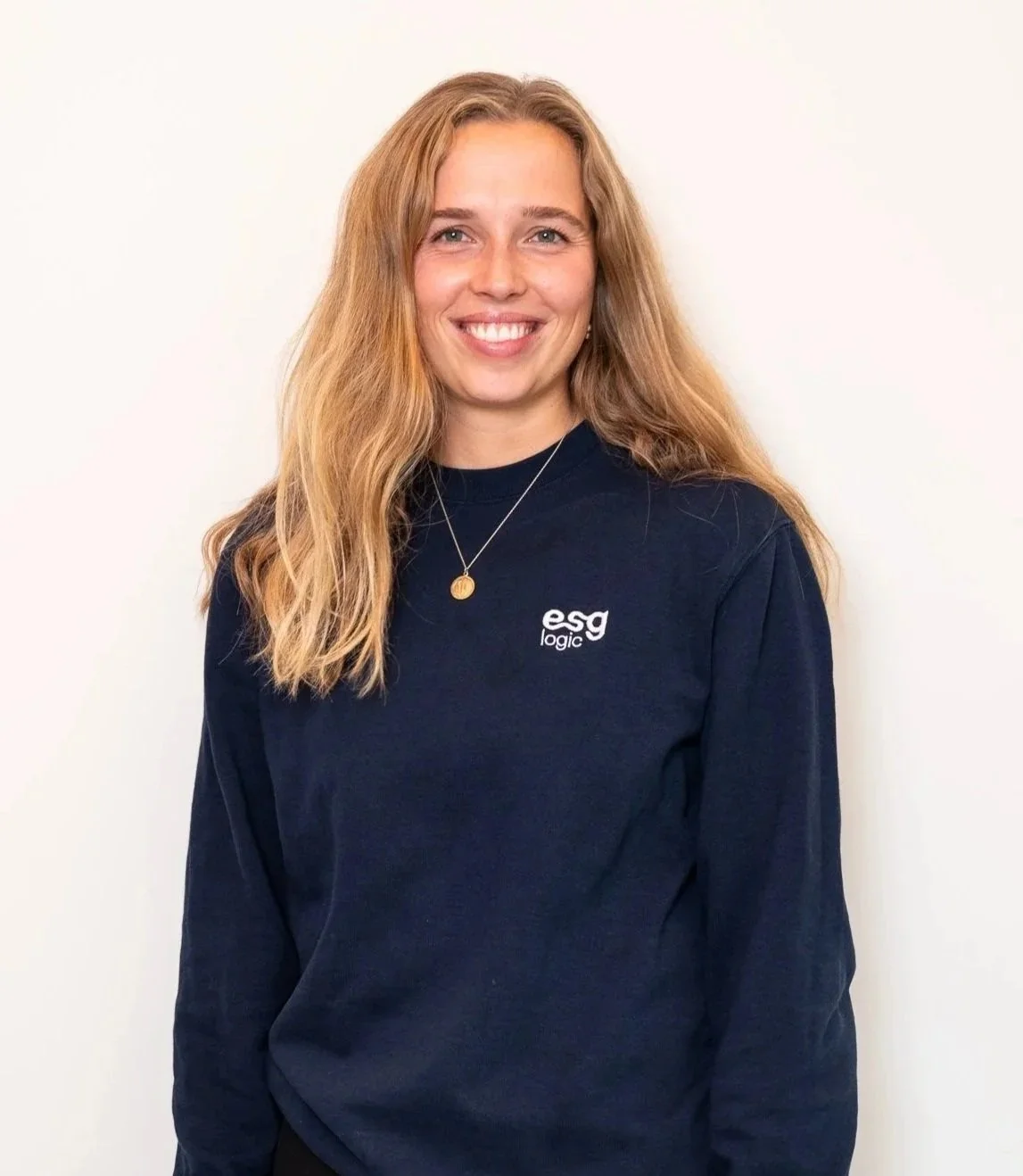 A young woman with long, wavy blonde hair smiling and wearing a navy blue sweatshirt with 'esg logic' logo, standing against a plain white wall.