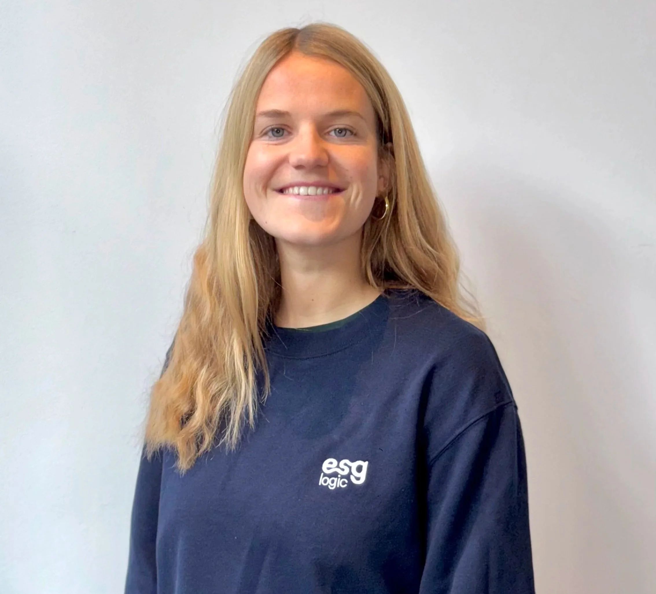 A woman with long blonde hair smiling, wearing a navy blue sweatshirt with 'esg logic' logo, standing against a plain light gray background.