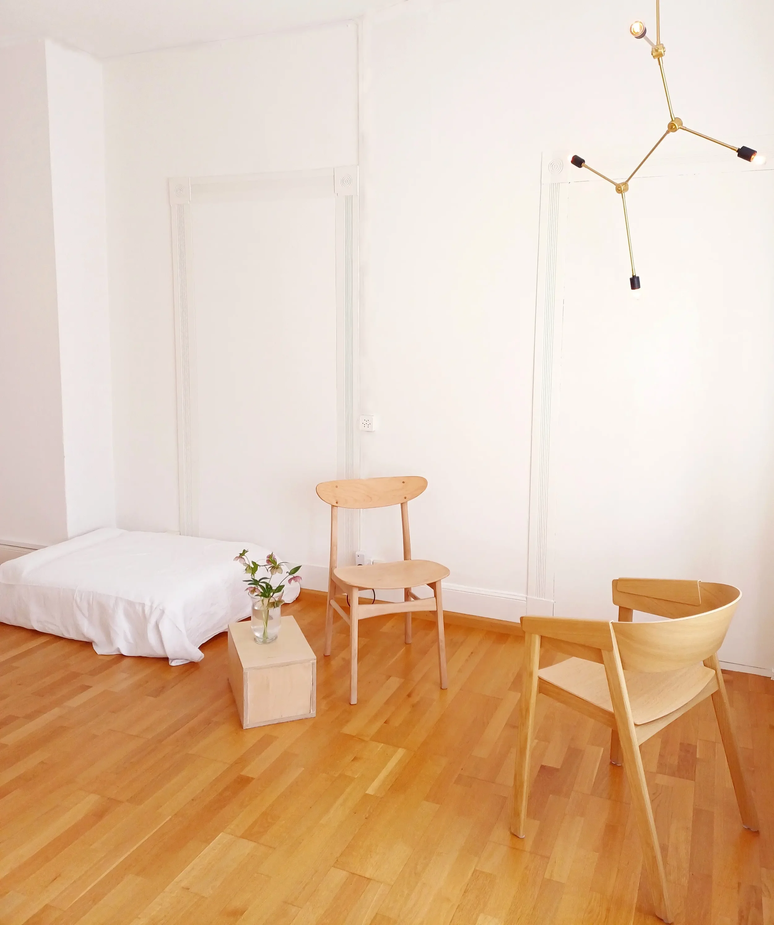 Therapy praxis with two light wood armchairs, a large cushion on the floor with a white linen sheet, a wooden shelf with a vase of flowers and pendant lighting.