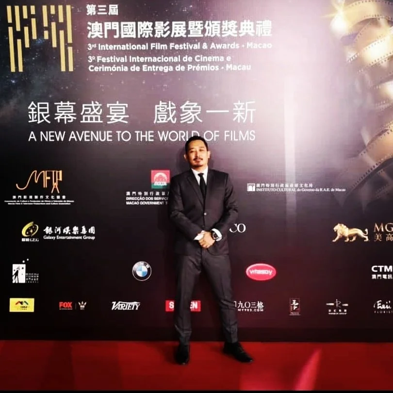 A man in a black suit standing in front of a large banner for the 3rd International Film Festival & Awards in Macau. The banner features Chinese, Portuguese, and English text, along with various sponsor logos and the tagline "A New Avenue to the Worl