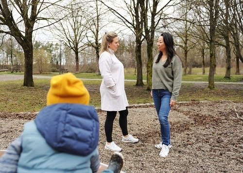 Zwei Frauen unterhalten sich auf dem Spielplatz, ein Kind sitzt vorne mit einer gelben Mütze und schaut zu