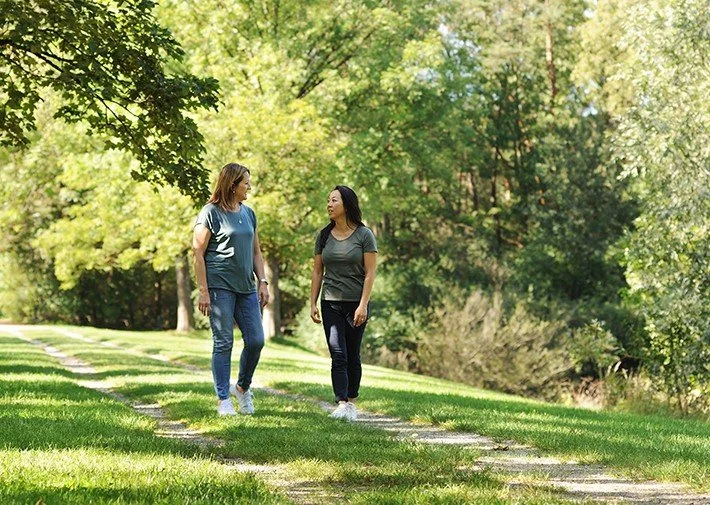 Zwei Frauen gehen in der Natur spazieren und schauen sich dabei an und unterhalten sich, eine hat schwarze Haare und die andere blonde in Gernlinden bei Psychotherapie im Gehen in der Natur