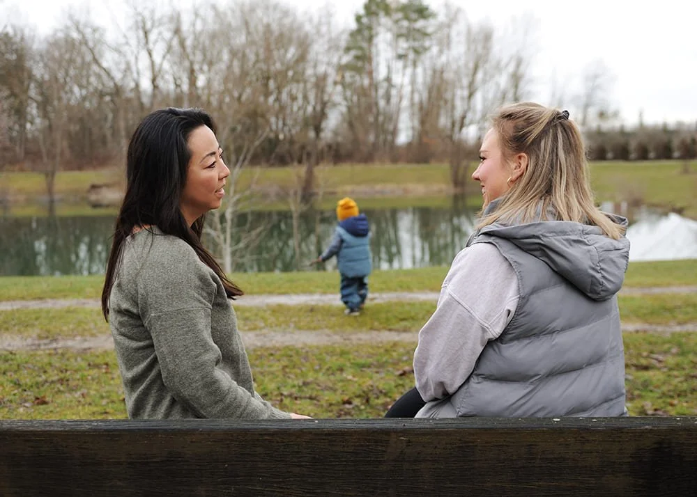 Psychotherapie in der Natur im Freien in Maisach bei Fürstenfeldbruck, zwei Frauen sitzen auf einer Parkbank und unterhalten sich, im Hintergrund spielt in Kind mit einem Stock und läuft Richtung See, eine hat schwarze und die andere blonde Haare
