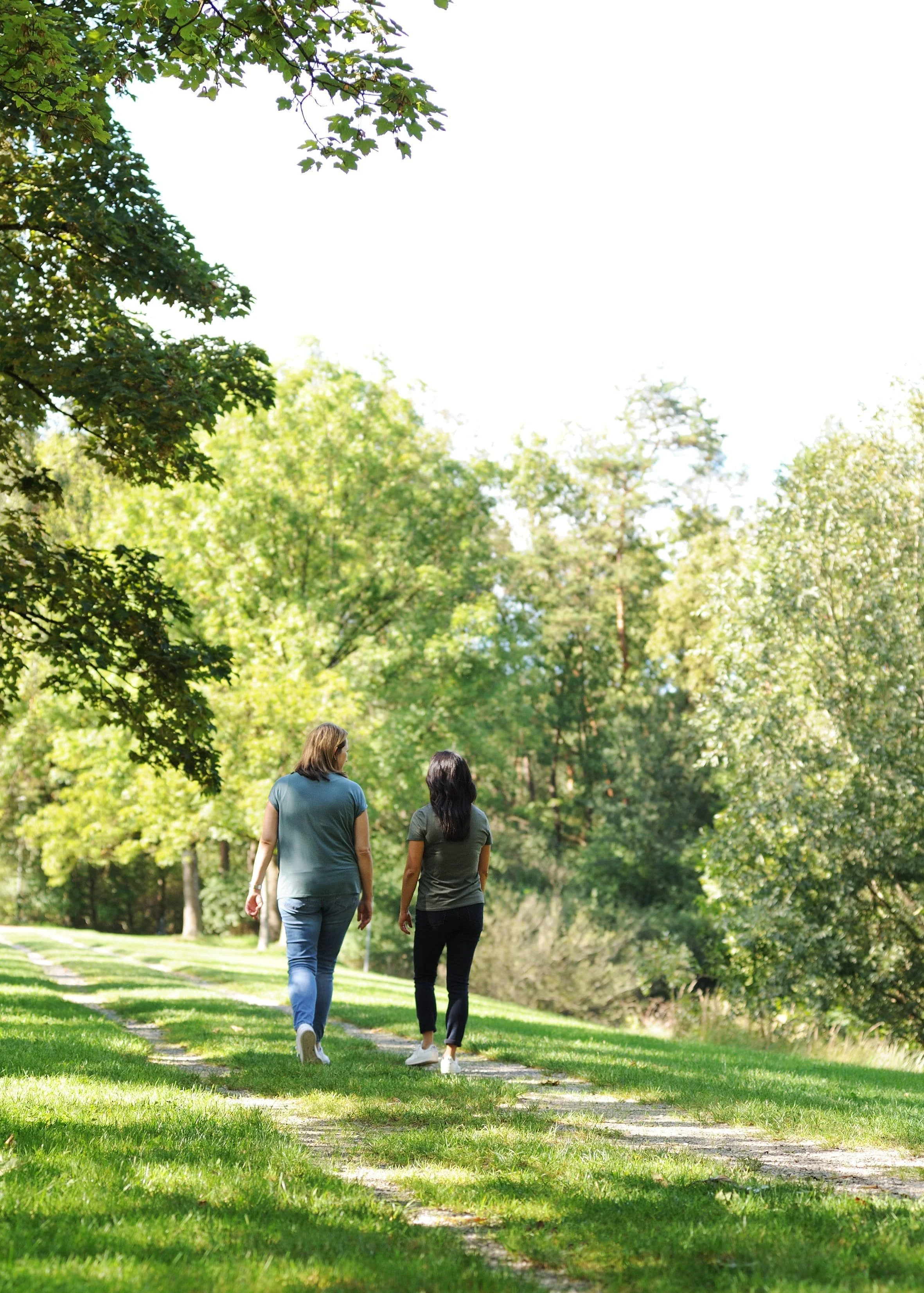 Zwei Frauen gehen in der Natur spazieren und sind von hinten zu sehen. Beide tragen eine lange Hose und ein T-Shirt