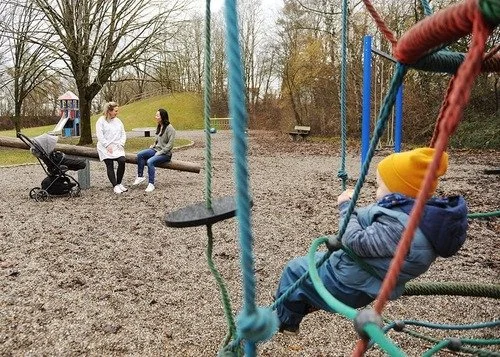 Ein Kind spielt im Vordergrund im Hintergrund sitzen zwei Frauen auf einer Wippe und unterhalten sich, neben einer Frau steht ein Kinderwagen, Psychotherapie im Landkreis Fürstenfeldbruck in Gernlinden auf dem Spielplatz