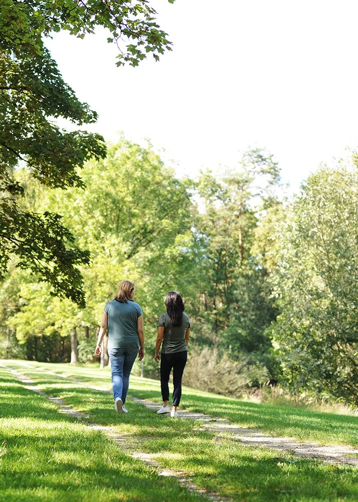 Es sind sind Frauen von hinten beim Spazierngehen ein der Natur zu sehen, eine hat blonde Haare und die andere schwarze Haare, beide tragen eine Jeans und ein grünes T-Shirt