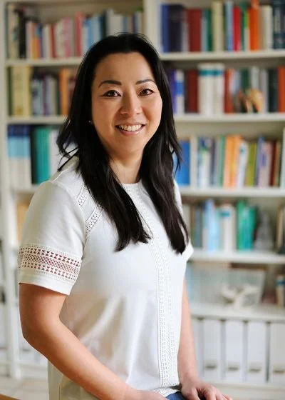 Chy-Eun Sohn, Heilpraktikerin für Psychotherapie in Maiasch im Landkreis Fürstenfeldbruck sitzt lächelnd vor einem Bücherregal, sie hat eine weiße kurze Bluse an und schwarze lange Haare
