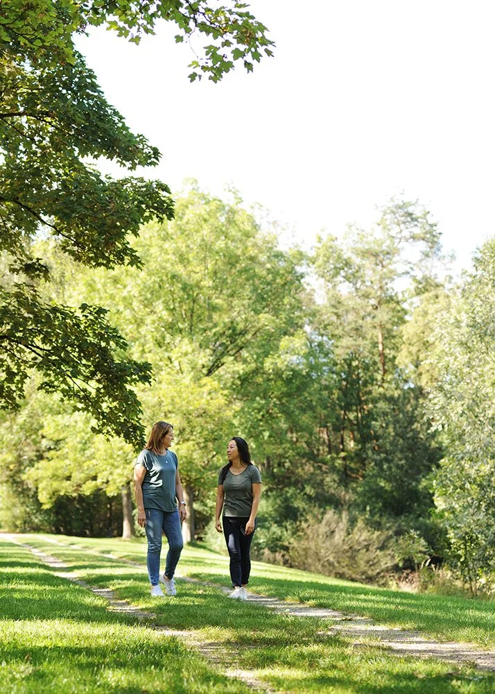 Chy-Eun Sohn führt ein Therapiegespräch am Gernlindner See in der Natur, sie hat lange schwarze Haare und die andere Frau hat blinde schulterlange Haare. Die Frauen schauen sich an und unterhalten sich, tragen grüne T-Shirts und Jeanshosen