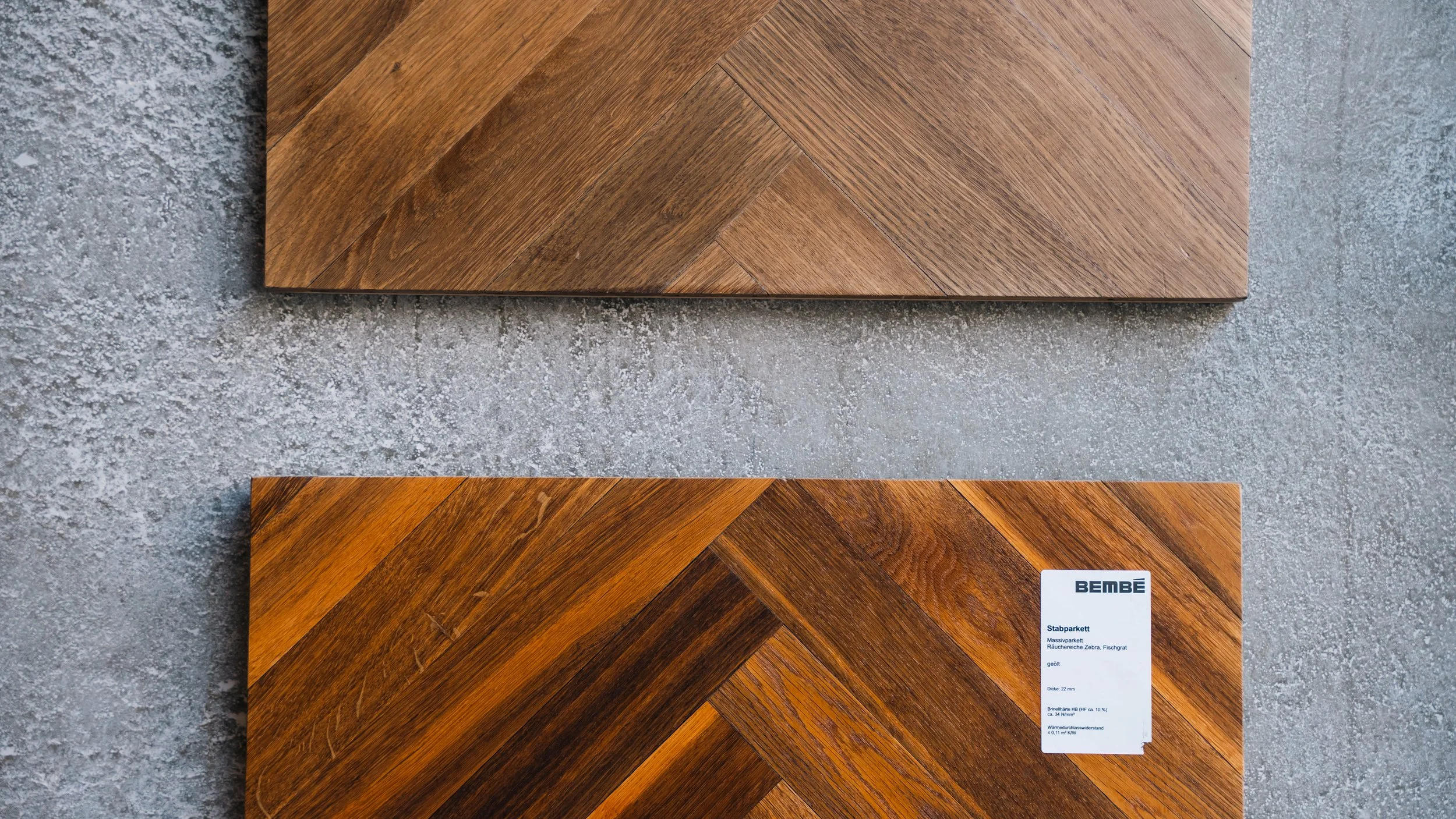 Two wooden parquet flooring tiles with a herringbone pattern on a gray textured surface. The tile at the bottom has a white label with blue text.
