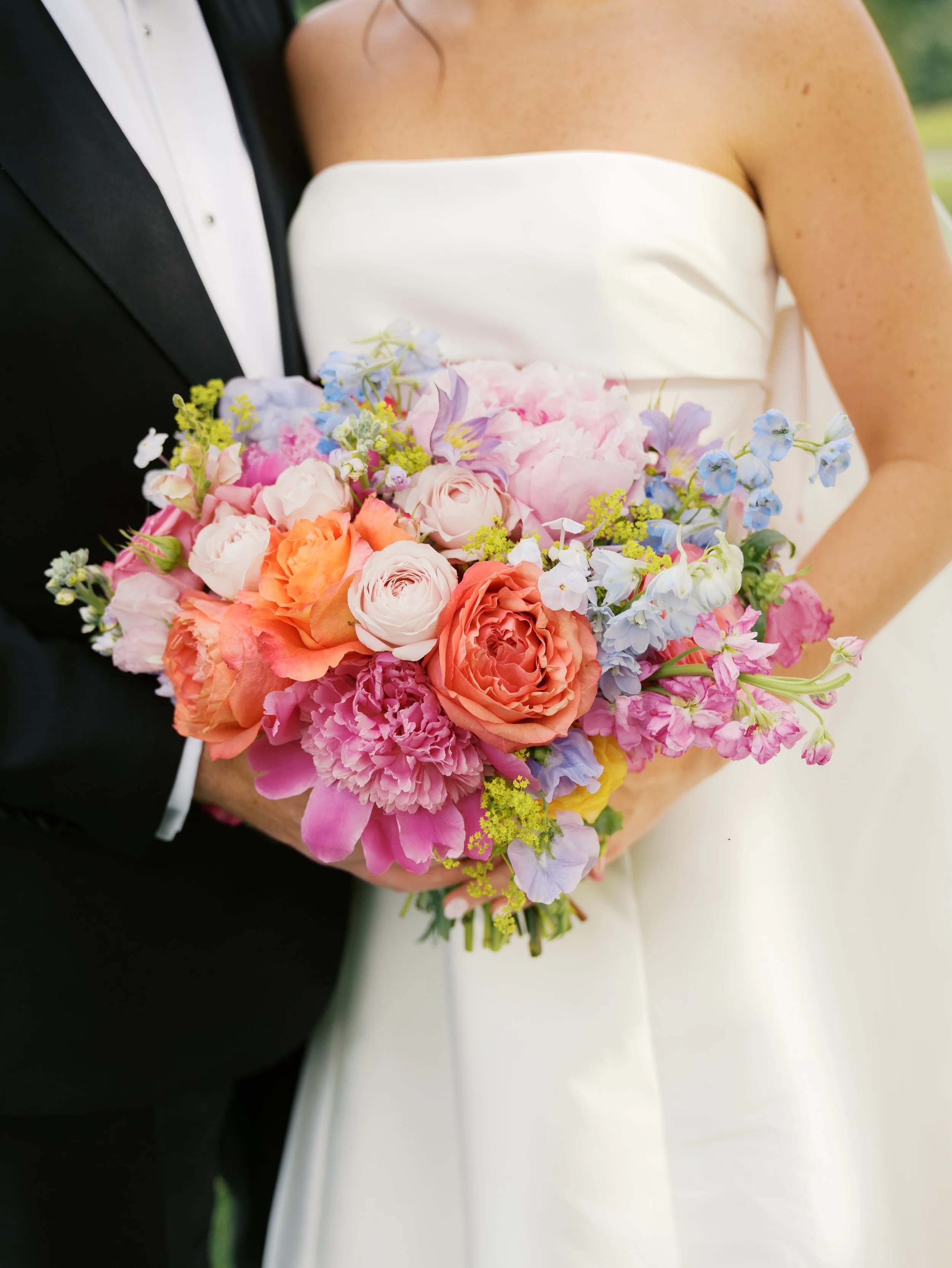 Close-up of a bride holding a colorful bouquet of flowers, wearing a strapless white wedding gown and standing next to a groom in a black tuxedo.